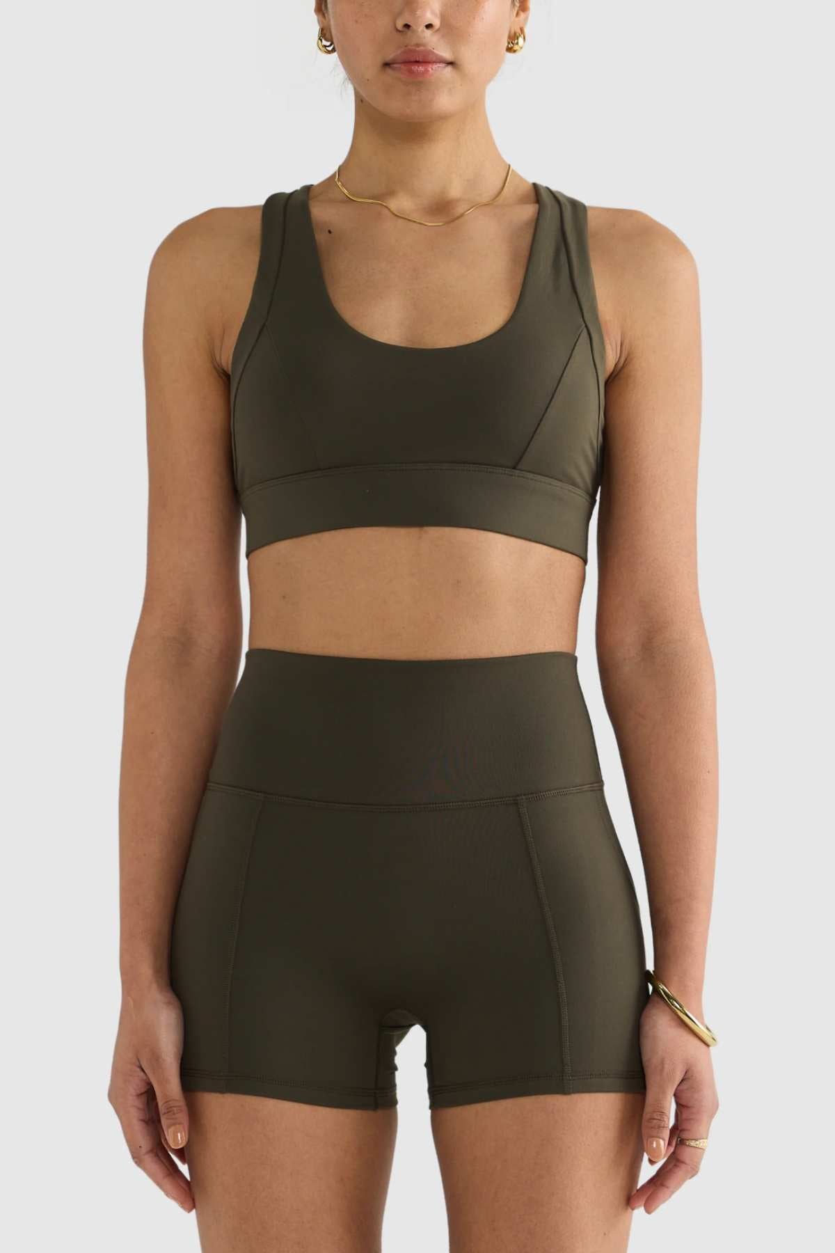 A person wearing the ORTC Eva Crop Top in olive green with matching high-waisted shorts, both crafted from recycled polyester, stands against a plain white background. They accessorize with a necklace and bracelet, completing the look with style and eco-consciousness.