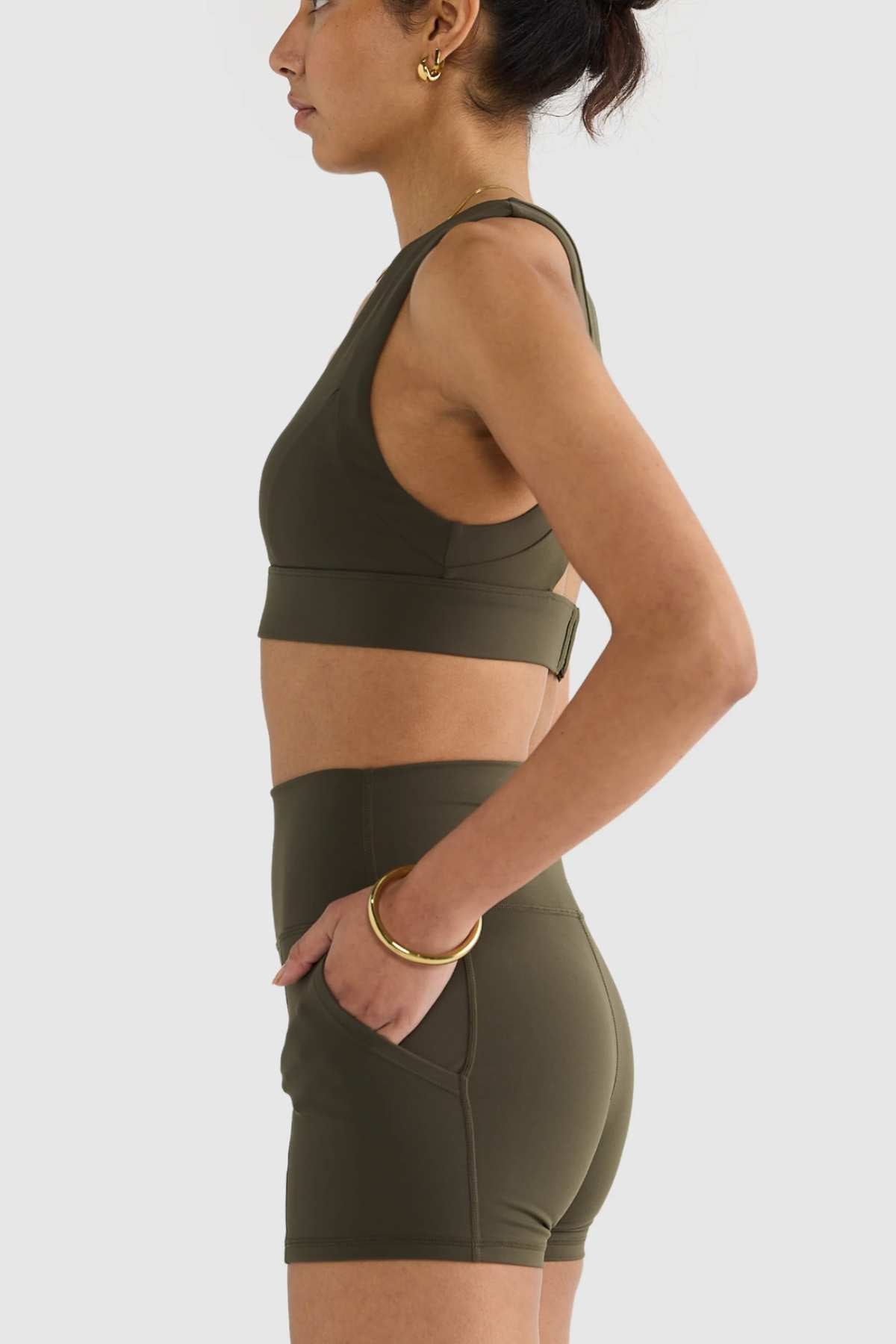 A person in profile is styled in the ORTC Eva Crop Top, a fashionable olive green piece paired with high-waisted shorts. The look is enhanced by gold hoop earrings and a matching bracelet, with one hand casually placed in their shorts pocket against a simple backdrop. Made from recycled polyester, the outfit provides firm support and sustainability.