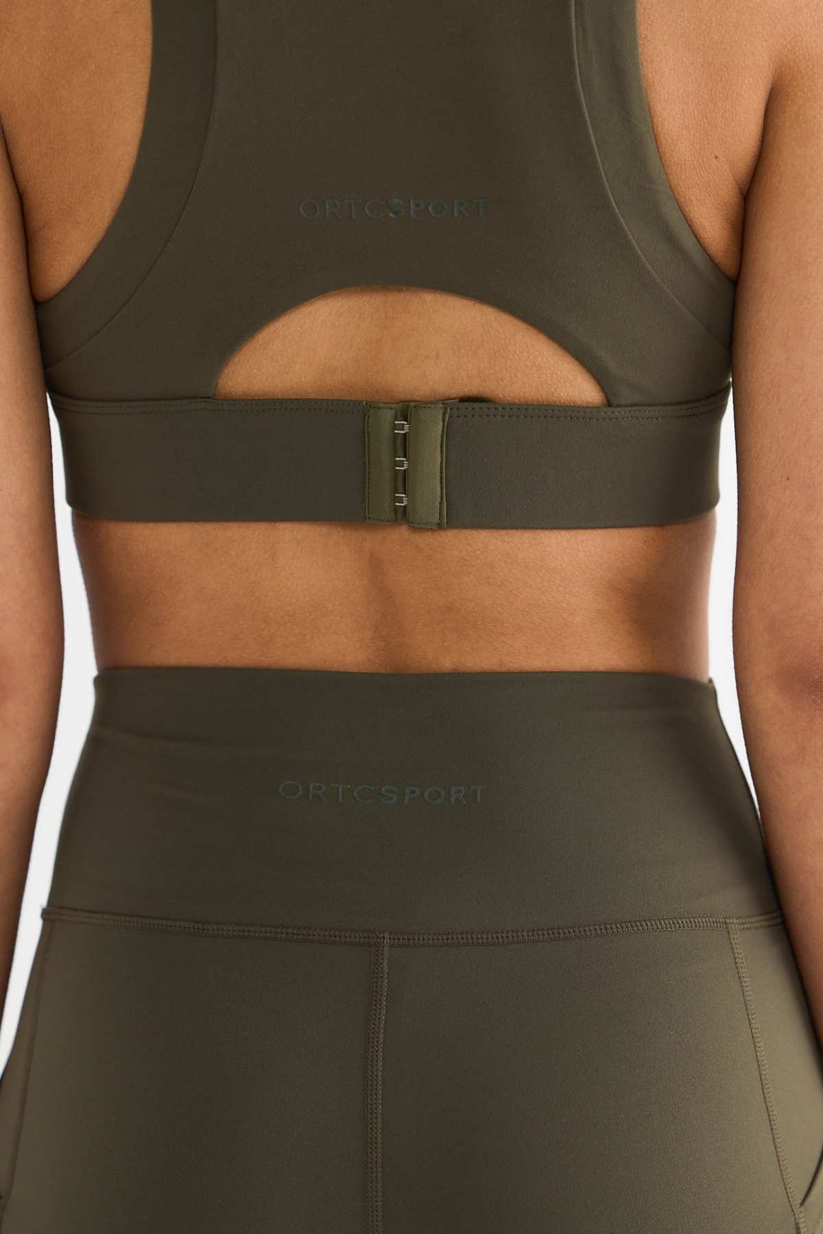 A person wearing a matching olive green ORTC Sport set, which includes the Eva Crop Top and leggings made from recycled polyester. The firmly supportive crop top features a hook clasp and a small cutout on the back, with the brand name visible on both pieces.
