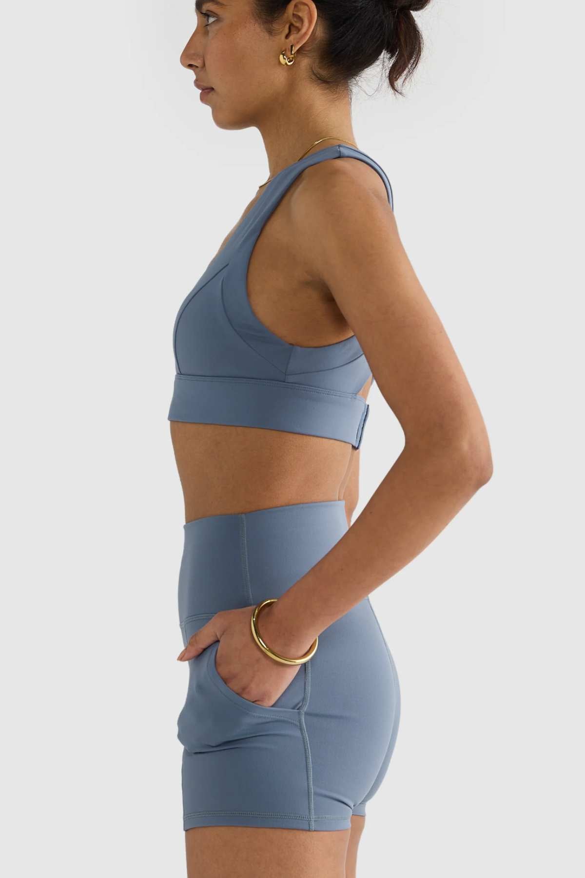 A person is shown in profile wearing the ORTC Eva Crop Top, paired with shorts. They accessorize with a gold hoop earring and bracelet, and their hair is styled in a bun. The plain white background highlights the top's durable fabric that offers firm support.