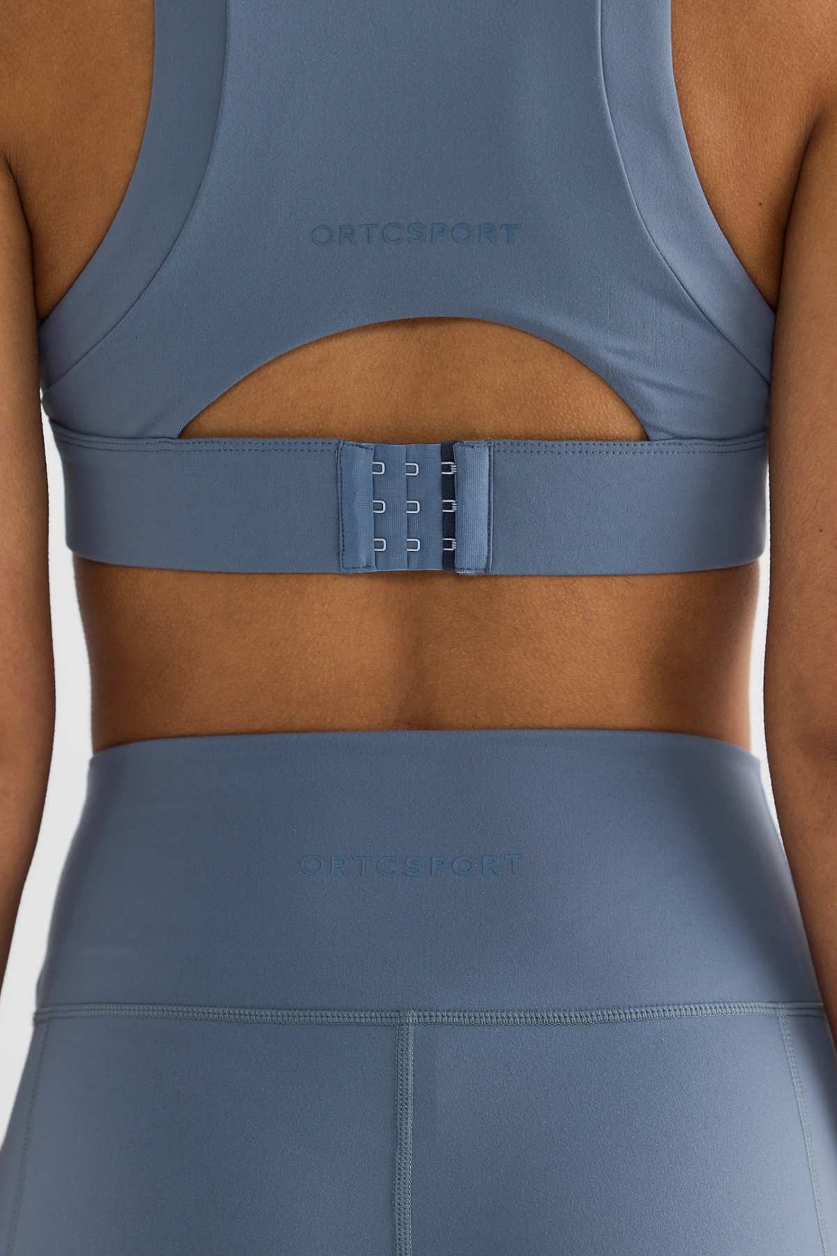 A person dressed in the ORTC Eva Crop Top sports bra along with matching blue leggings. The picture emphasizes the solid support and long-lasting fabric, showing off the clasp of the sports bra and the high waistband of the leggings, with the "ORTCSPORT" brand logo prominently displayed on both pieces.