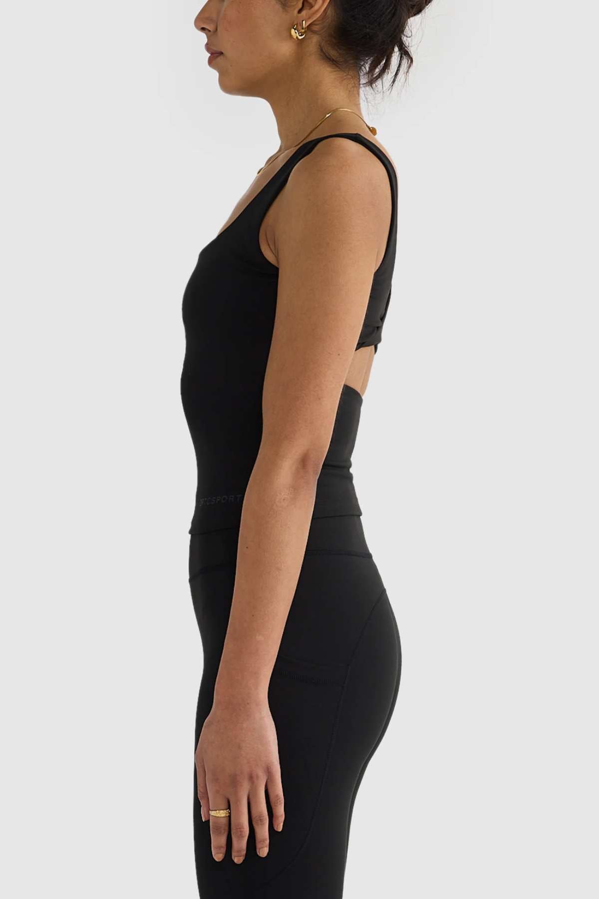 A person with dark hair stands in profile against a plain background, wearing the ORTC Eva Twist Back Reversible Top and leggings. They have hoop earrings and a ring on their finger, showcasing the top's dual-wear design that seamlessly merges style with functionality.