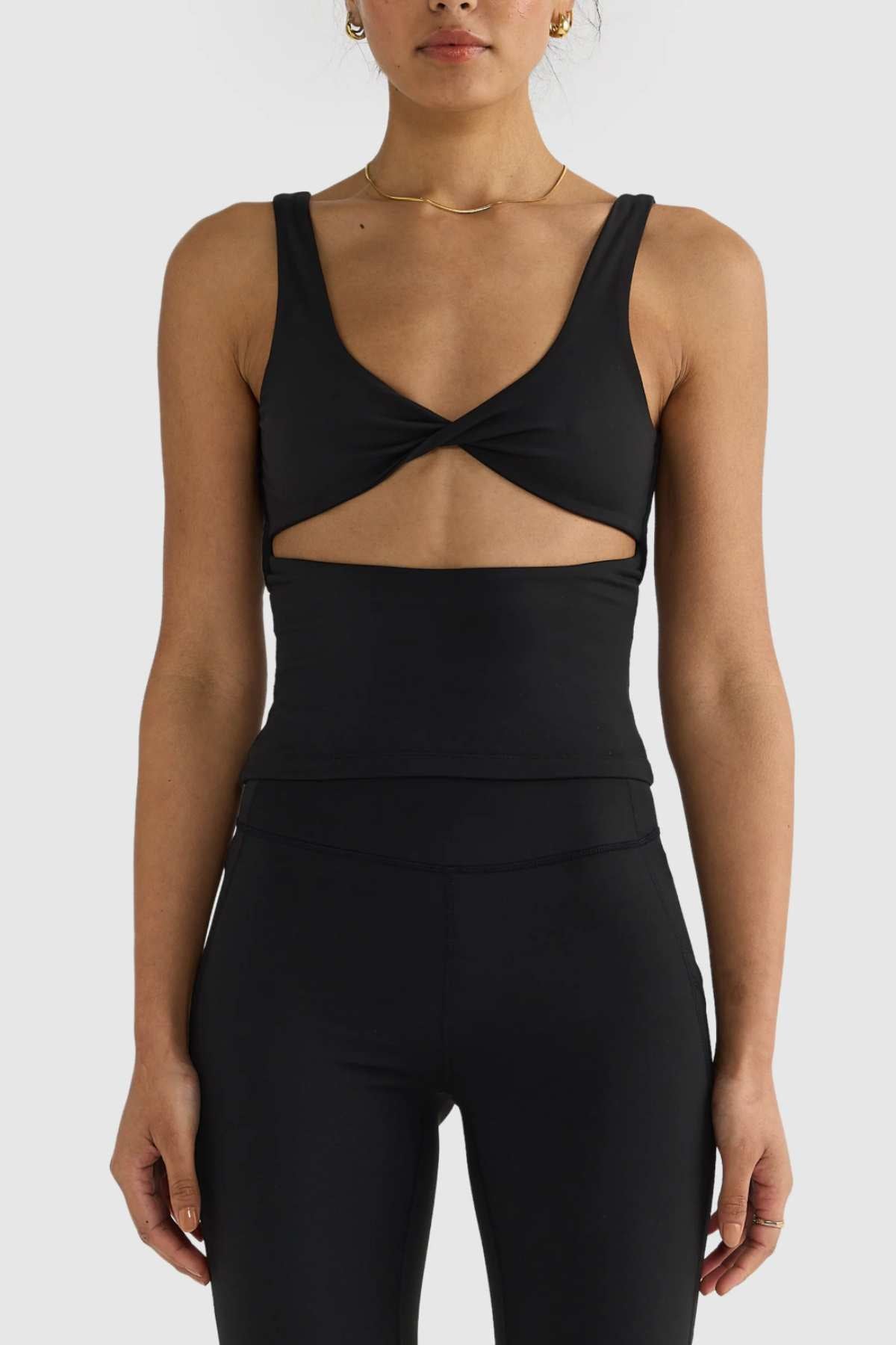 A person wearing the ORTC Eva Twist Back Reversible Top, a sleek black sleeveless piece with a twisted front design, paired with high-waisted leggings stands confidently against a plain gray background. This versatile top offers dual-wear design possibilities while maintaining an effortlessly chic look.