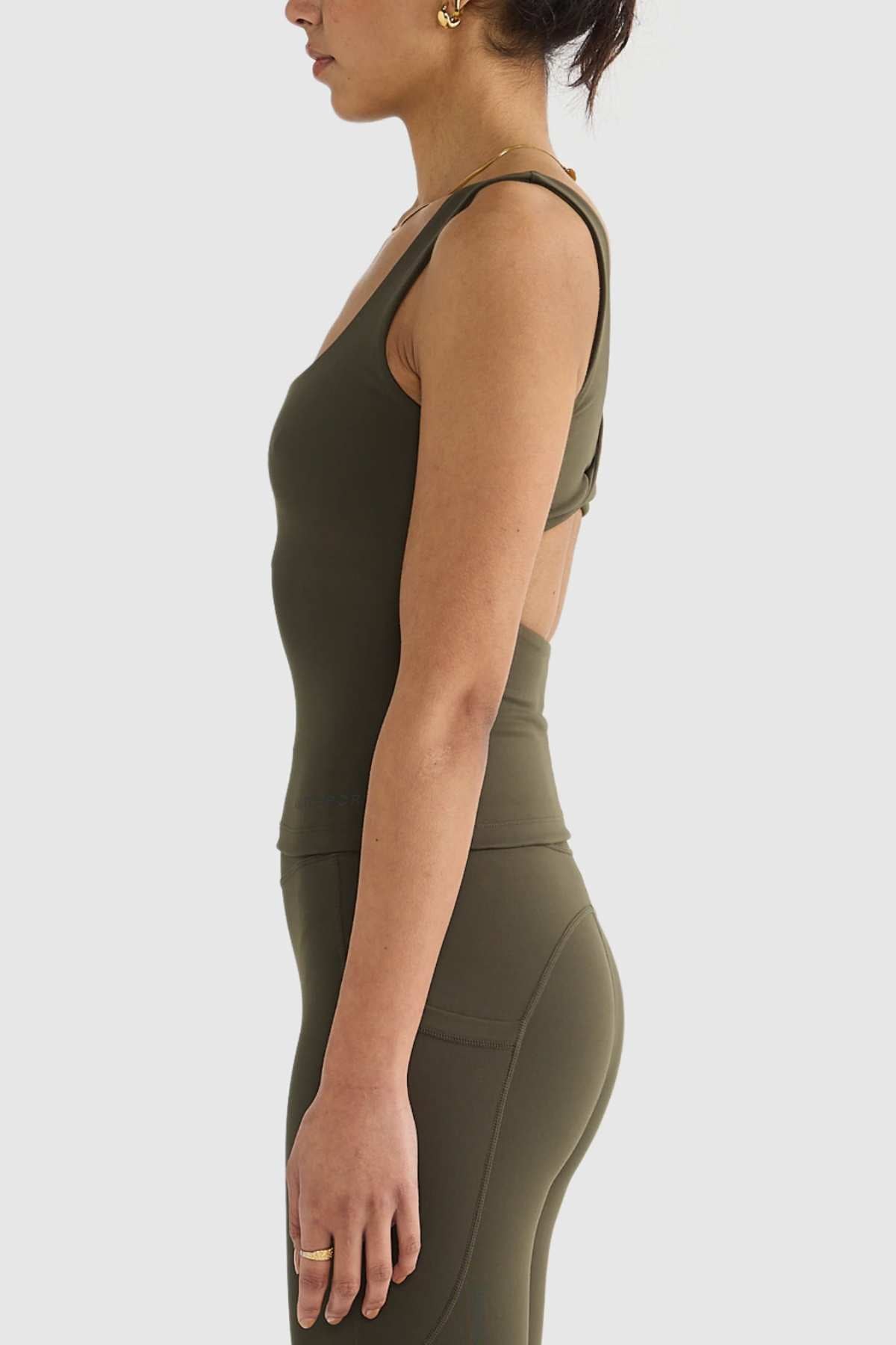An individual dressed in an olive green activewear outfit is pictured in profile against a light background. They are wearing the ORTC Eva Twist Back Reversible Top, crafted from ultra-soft double-brushed fabric with a crossover back design, along with matching leggings. Their accessories consist of earrings and a ring.