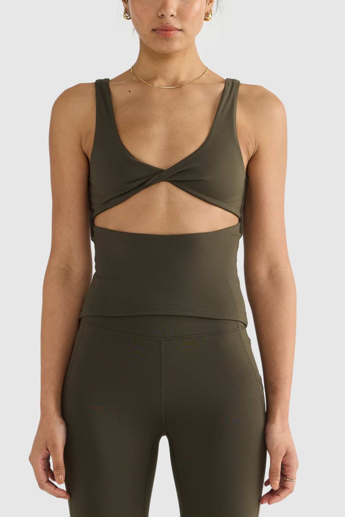 The woman is wearing an ORTC Eva Twist Back Reversible Top in dark green, highlighting its versatile dual-wear design with a twist front cutout. She completes her look with matching high-waisted leggings, both crafted from ultra-soft double-brushed fabric for maximum comfort.