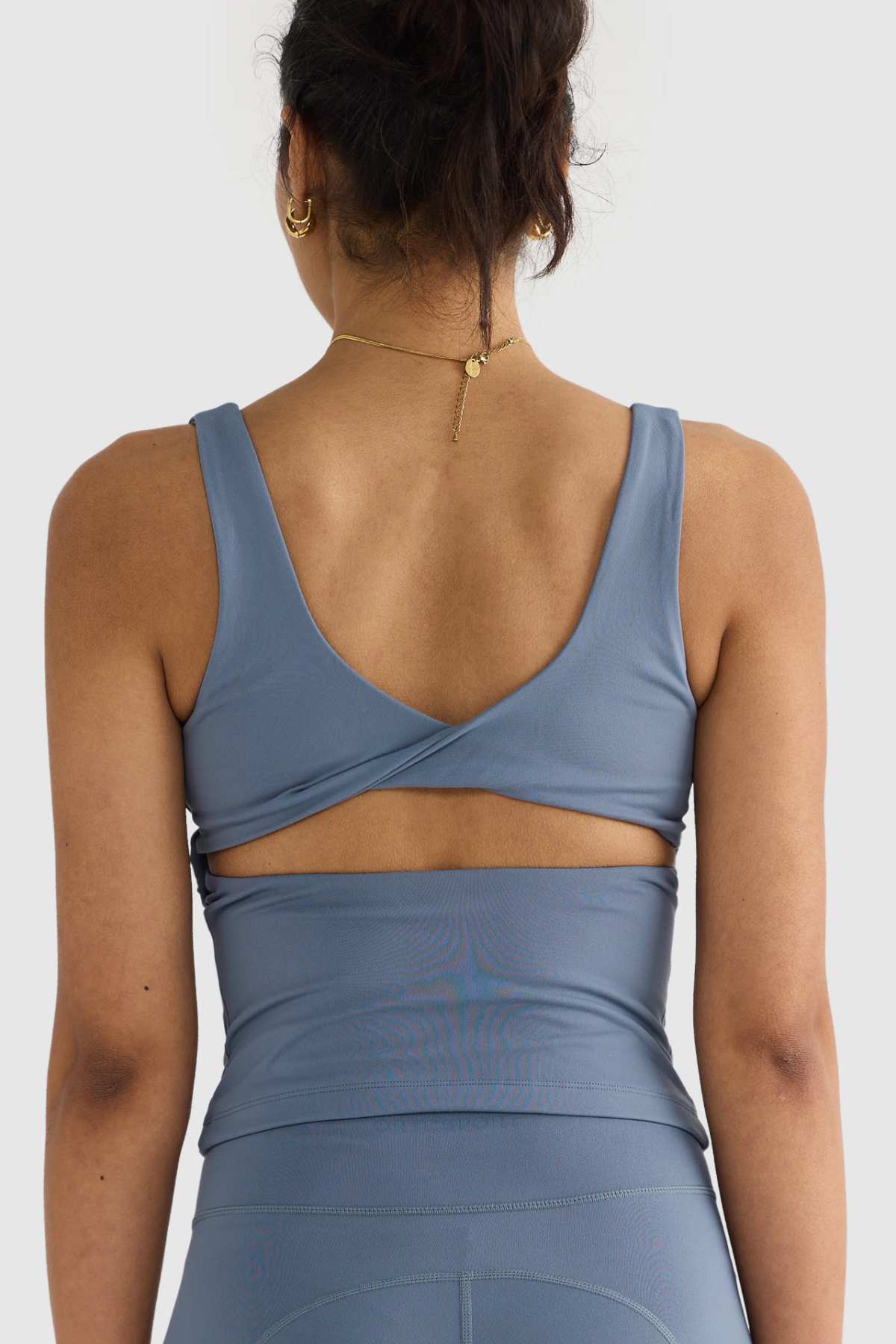 A woman is shown from the back wearing a blue ORTC Eva Twist Back Reversible Top with cross-back straps, paired with matching leggings. Her hair is tied up, and she accessorizes with a necklace featuring a small pendant. The double-brushed fabric enhances the sleek appearance against a simple light gray background.