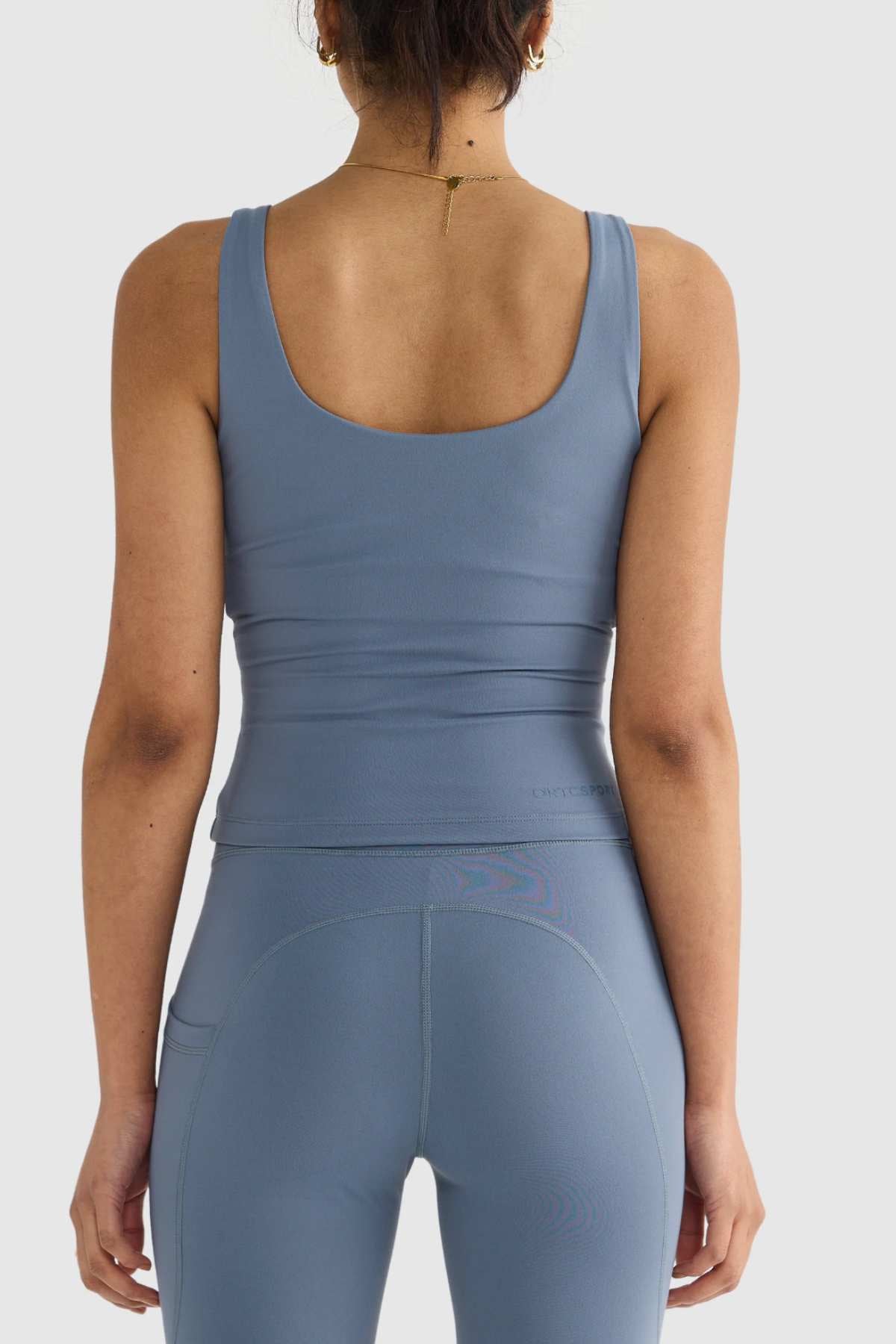 A person wearing a coordinated blue athletic tank top and leggings, featuring the ORTC Eva Twist Back Reversible Top design, stands with their back facing the camera. They have a thin gold chain necklace and their hair tied up, against a plain white background.
