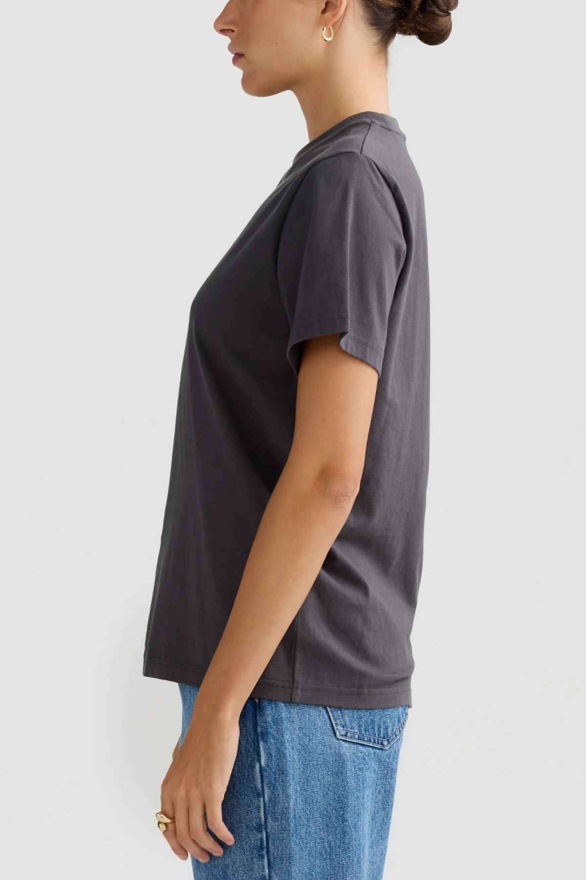 A woman is pictured in profile against a simple backdrop, dressed in a dark gray ORTC Everyday T-Shirt paired with blue jeans. Crafted from soft mid-weight cotton, the shirt provides a relaxed fit. Her hair is styled up, she wears small hoop earrings, and her hands are casually placed at her sides.