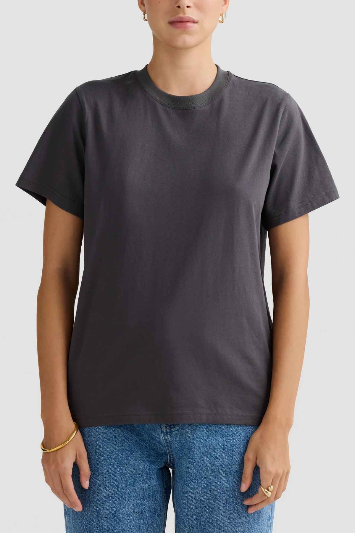 A person wearing a relaxed fit ORTC Everyday T-Shirt in plain dark gray pairs it with blue jeans while standing against a light gray background. Their hands rest casually by their sides, highlighting the gold hoop earrings and bracelets. Made from soft mid-weight cotton, this ensemble exudes an effortlessly casual vibe.