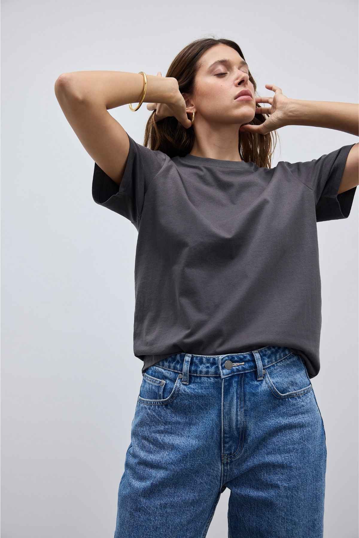 A person with long hair is dressed in an ORTC Everyday T-Shirt and blue jeans. The shirt, in a relaxed fit, adds to their style as they gently touch their ears with eyes closed. Made from soft mid-weight cotton, the dark gray T-shirt contrasts beautifully against the plain, light-colored background.