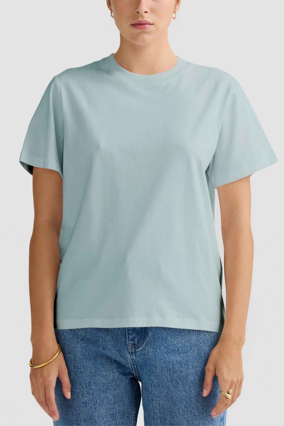 A person clad in the ORTC Everyday T-Shirt in light blue and relaxed blue jeans poses against a simple white backdrop, radiating casual elegance. Their short hair is perfectly set off by gold hoop earrings and a gold bracelet.
