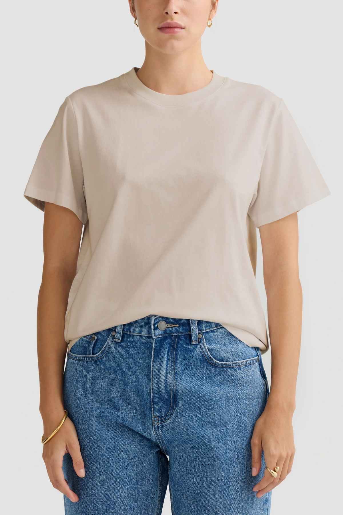 A person wearing an ORTC Everyday T-Shirt crafted from mid-weight cotton in beige, paired with blue jeans, stands against a plain background. They have short hair and are accessorized with gold hoop earrings and a bracelet, showcasing a relaxed fit and style.