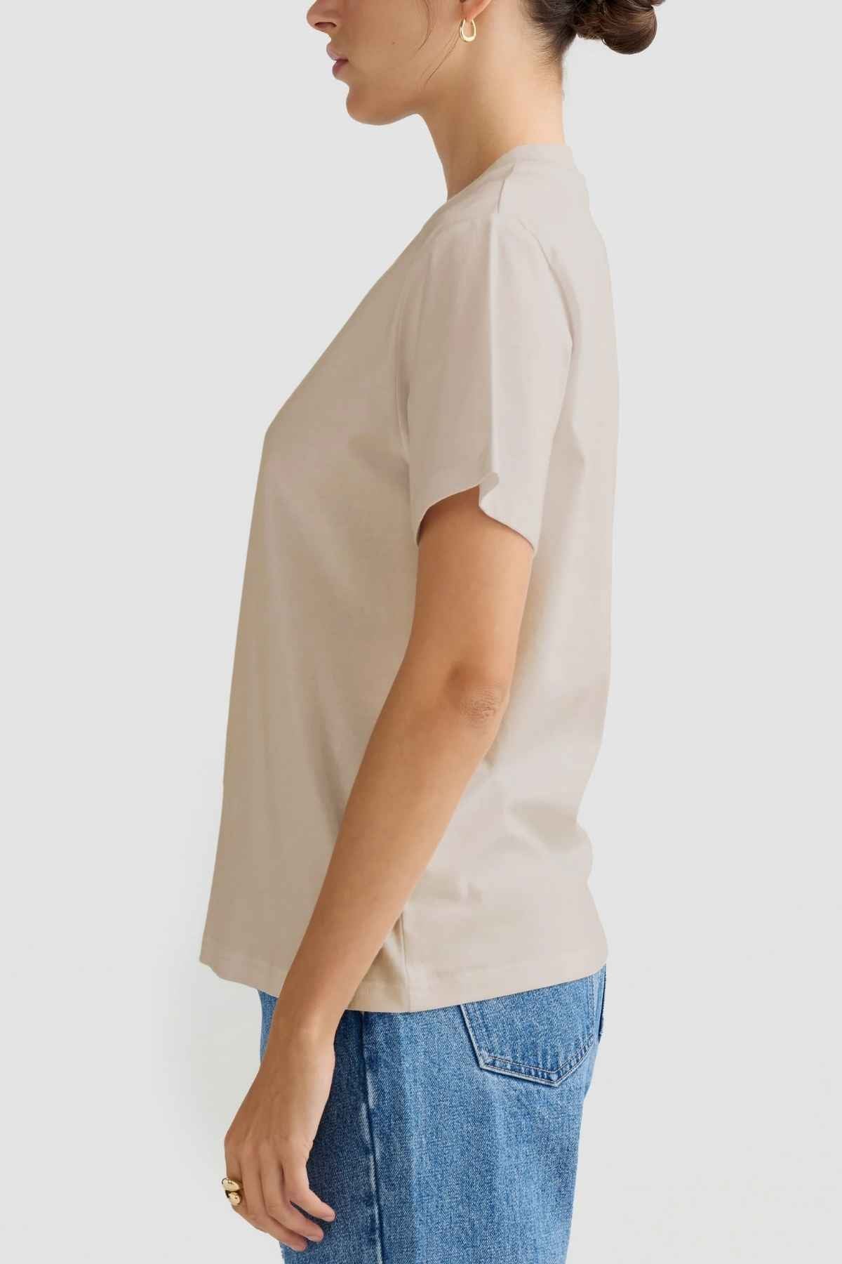 A person wearing an ORTC Everyday T-Shirt in a relaxed fit beige, crafted from mid-weight cotton, pairs it with blue jeans as they stand in profile against a light gray background. Their short hair is pulled back, and they sport a hoop earring along with a ring on their right hand, perfectly capturing the casual essence of the Everyday T-Shirt style.