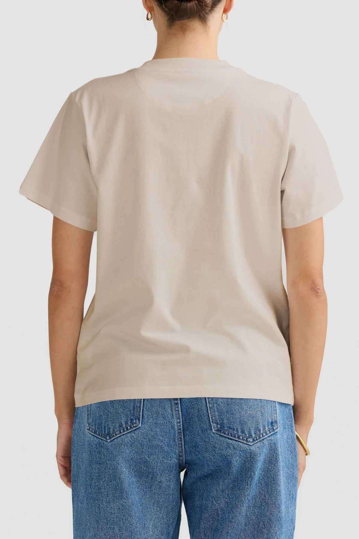 A person is photographed from the back wearing a relaxed fit, ORTC Everyday T-Shirt in beige paired with blue jeans. The mid-weight cotton tee stands out against the plain white background.
