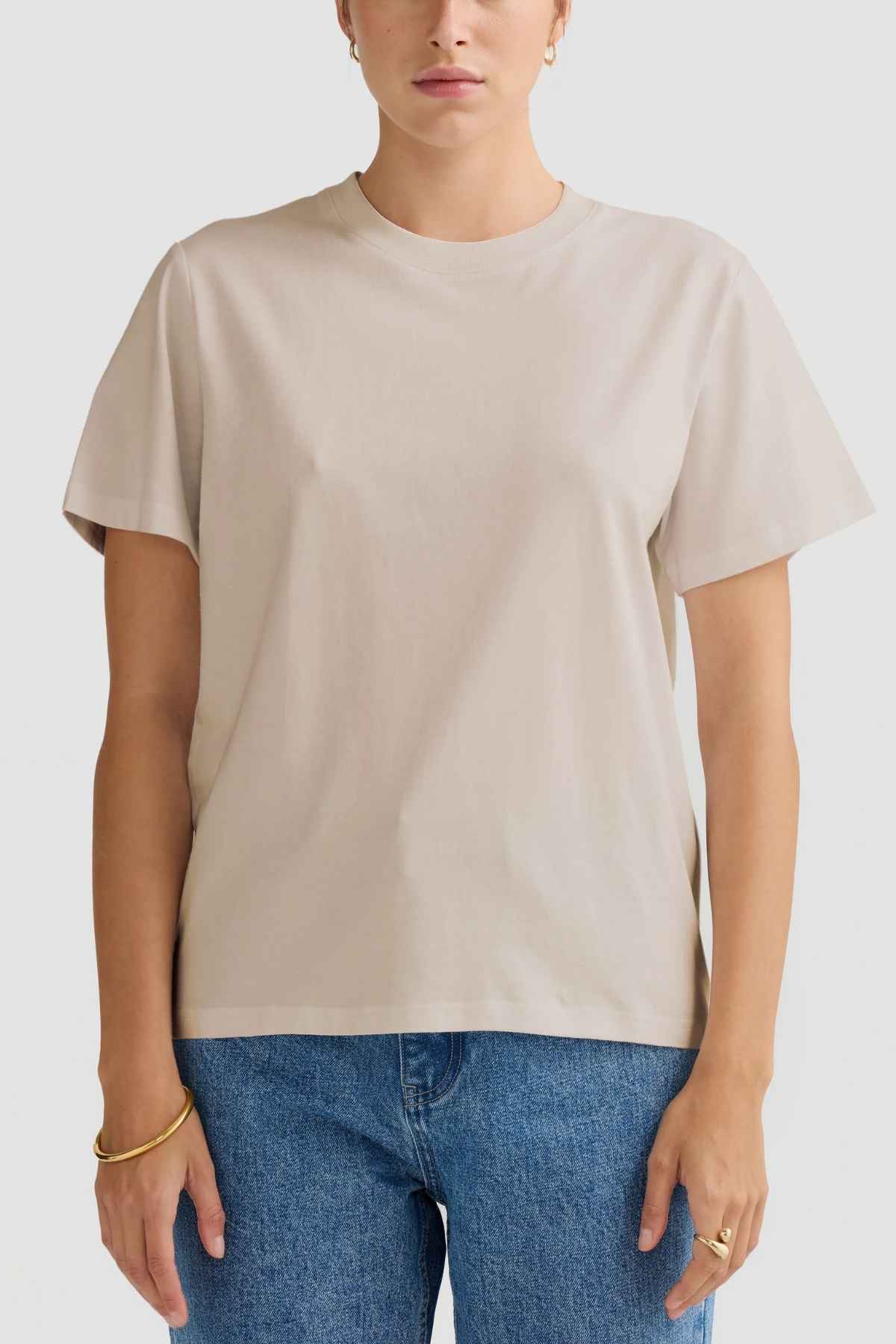 A person wearing an ORTC Everyday T-Shirt, made from mid-weight cotton, and blue jeans stands against a light gray background. The relaxed fit enhances the casual look, with gold bracelets and small hoop earrings providing an elegant touch.