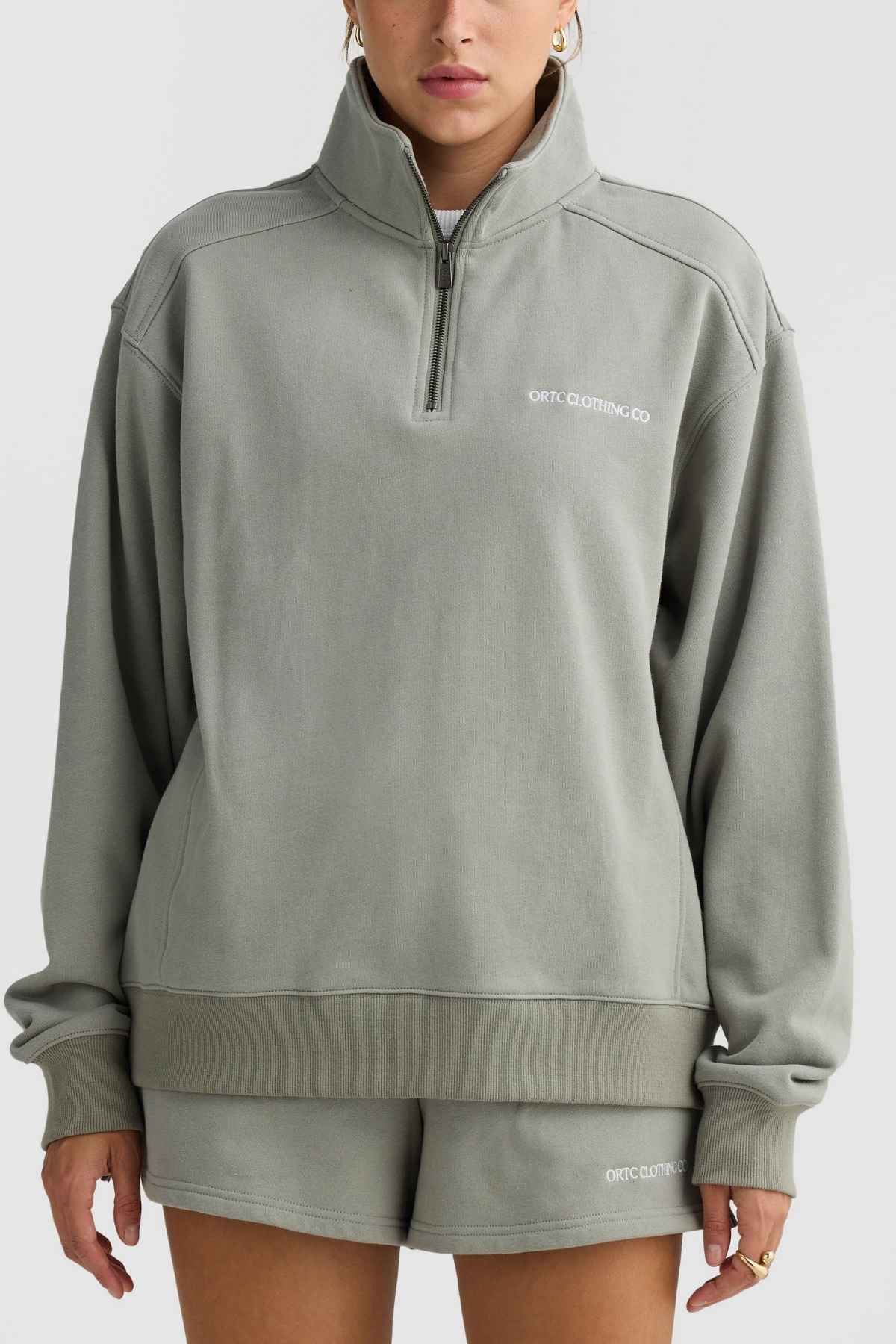 An individual dressed in the ORTC Lena Quarter Zip, a light gray-green terry cotton sweatshirt paired with matching shorts. The sweatshirt features a high collar adorned with the "ORTC CLOTHING CO." branding, which also appears on the shorts. They are set against a plain gray backdrop.