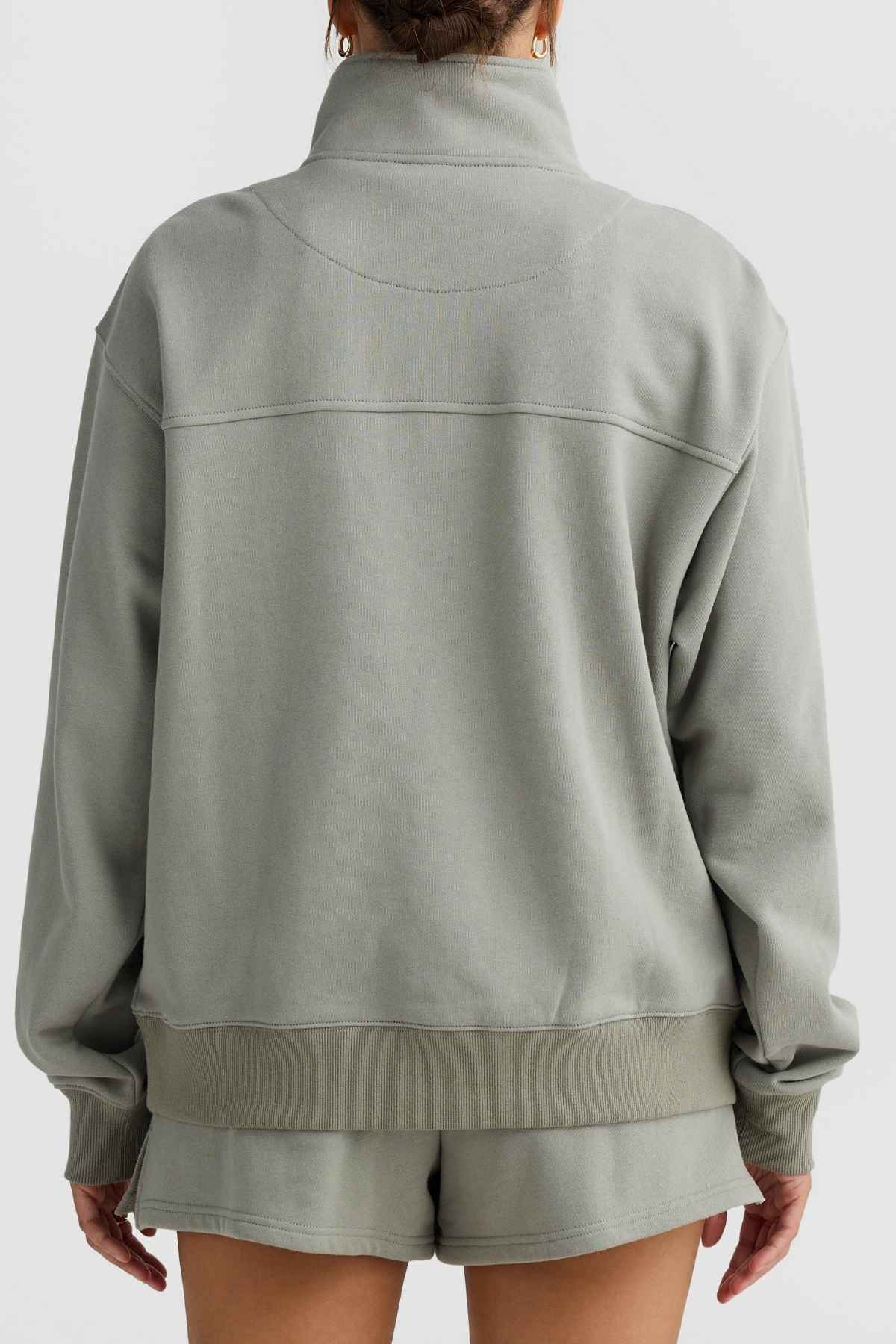 A person clad in the light gray ORTC Lena Quarter Zip sweatshirt and Terry Cotton shorts is facing away from the camera. The quarter zip features a high collar and relaxed fit, highlighted by visible seam details on the back. The background is plain and light-colored.