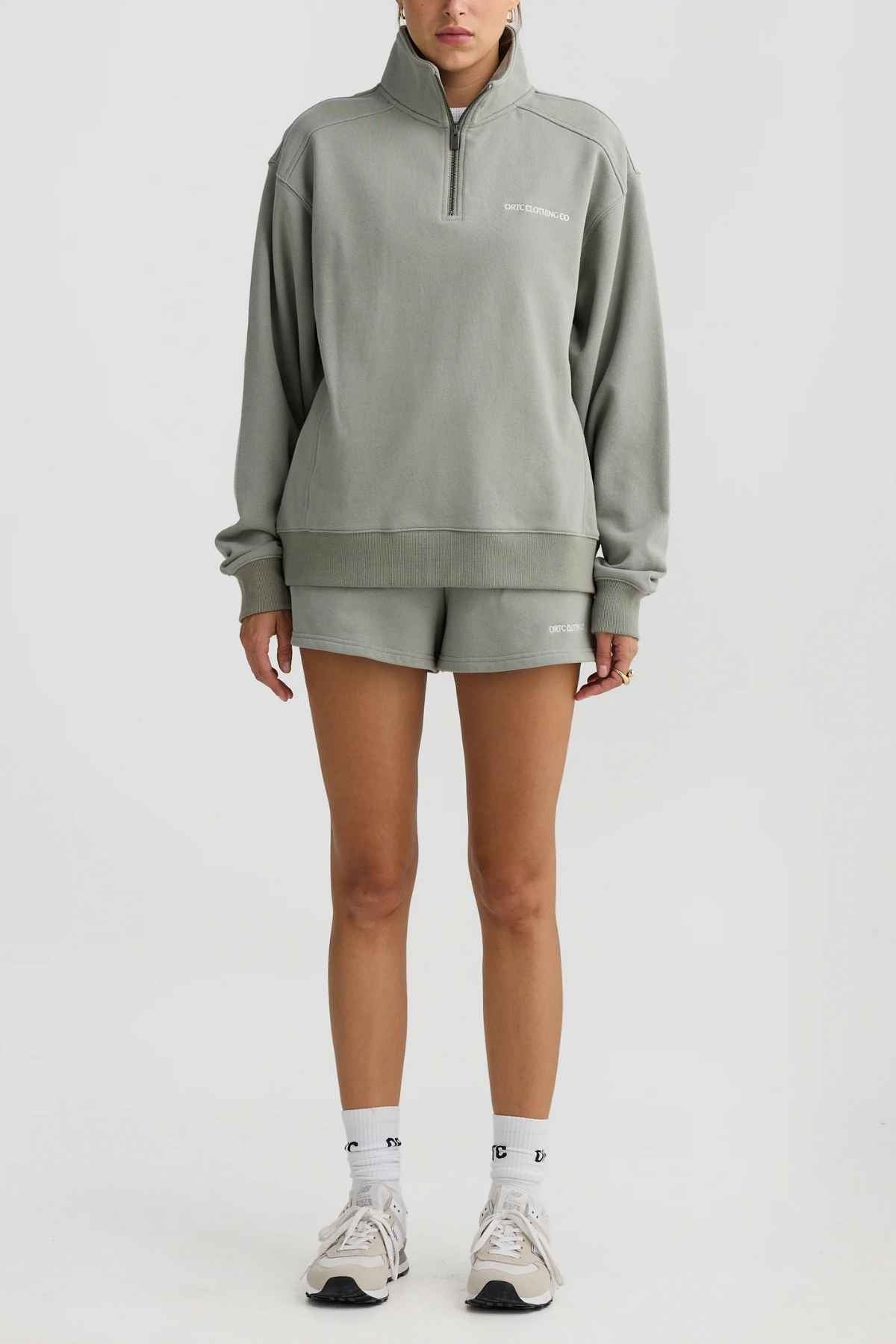 A person wearing a terry cotton ORTC Lena Quarter Zip sweatshirt and shorts set stands against a plain background. They sport white socks and sneakers. The quarter zip sweatshirt features subtle text on the left chest, adding a touch of style to their casual look.
