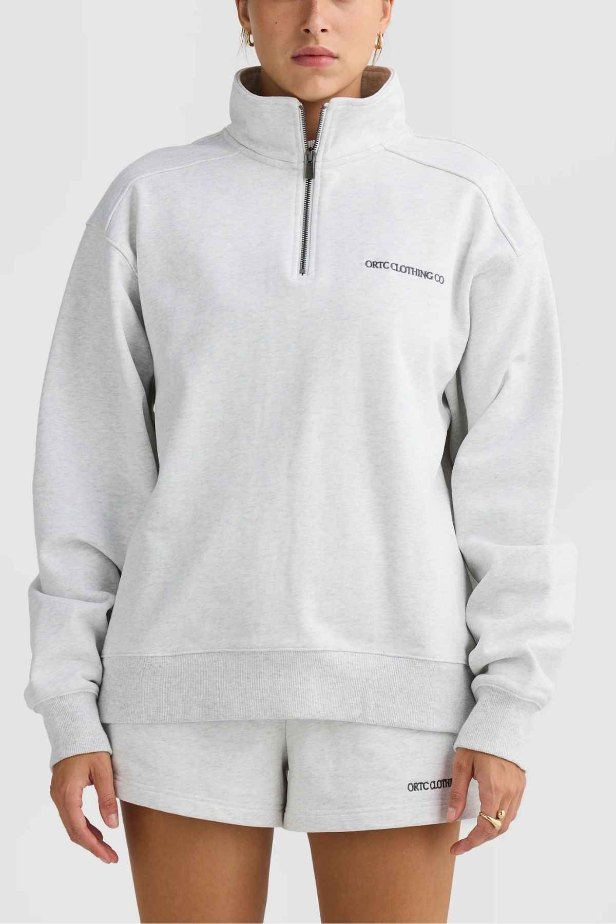 A person models the ORTC Lena Quarter Zip in light gray Terry Cotton, complemented by matching shorts. The ensemble is highlighted by an ORTC-branded metal zipper and a discreet logo on both the chest and shorts, set against a solid white background.