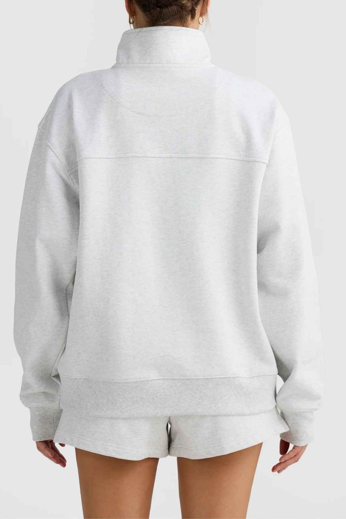 The individual is seen from the back, donning a light gray ensemble consisting of a sweatshirt and matching shorts. The ORTC Lena Quarter Zip stands out with its high collar and relaxed fit, accompanied by a branded metal zipper from ORTC. The backdrop is a simple, light gray shade.