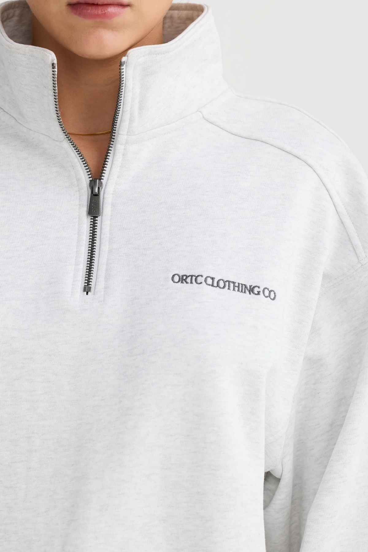 A person is wearing an ORTC Lena Quarter Zip in light gray, prominently displaying "ORTC CLOTHING CO" on the chest. Crafted from Terry Cotton, it includes a high collar and is accented with an ORTC branded metal zipper. The individual's hair is partially visible, but their face is largely out of frame.