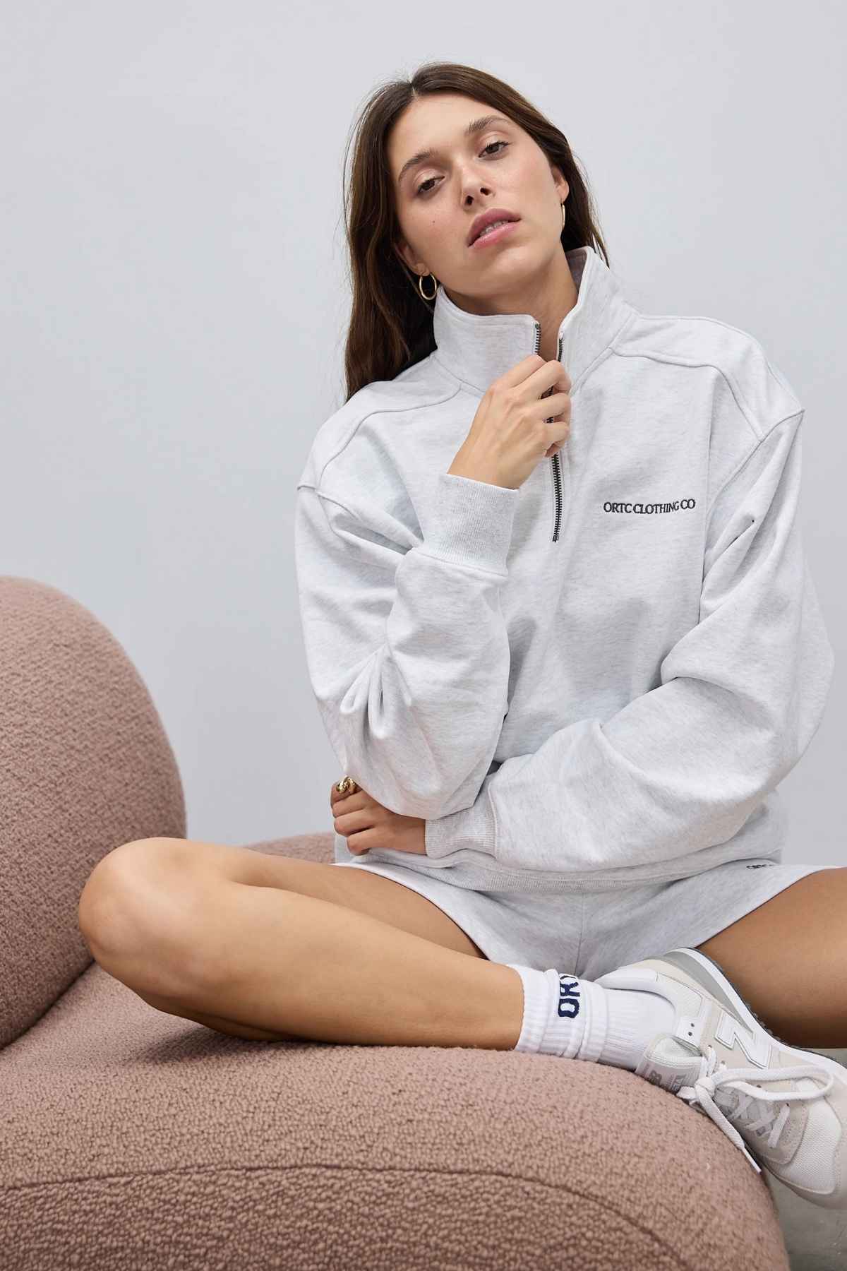 A person in an ORTC Lena Quarter Zip sweatshirt and matching shorts sits on a textured beige chair. The terry cotton fabric enhances their long hair, white socks, and sneakers, while the ORTC branded metal zipper provides a sleek detail against the plain, light-colored wall behind them.