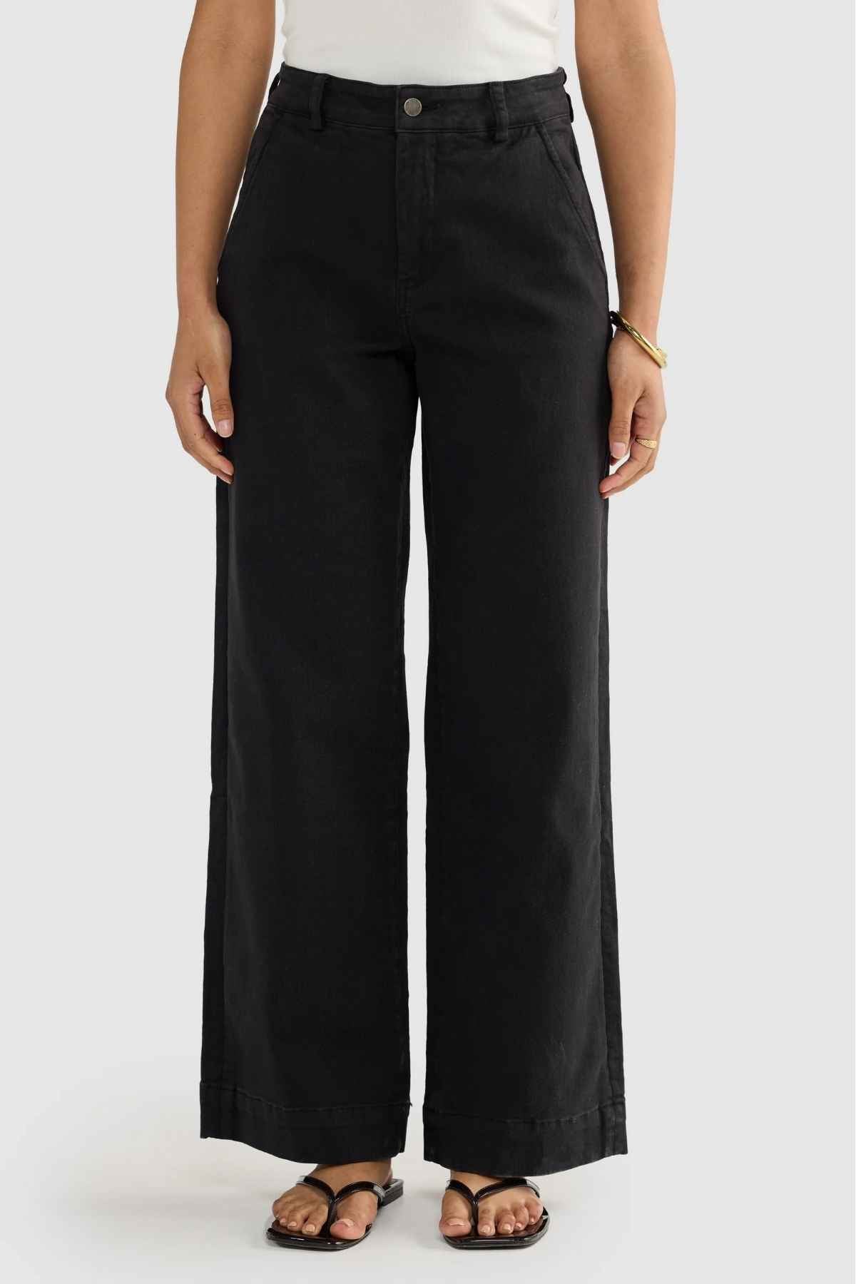 Wearing the relaxed-fit, high-waisted ORTC Lottie Relaxed Twill Pants with their wide-leg design, an individual effortlessly showcases style. Complemented by a basic white top and black sandals set against a light gray backdrop, the look is finished with a gold bangle adorning their left wrist.