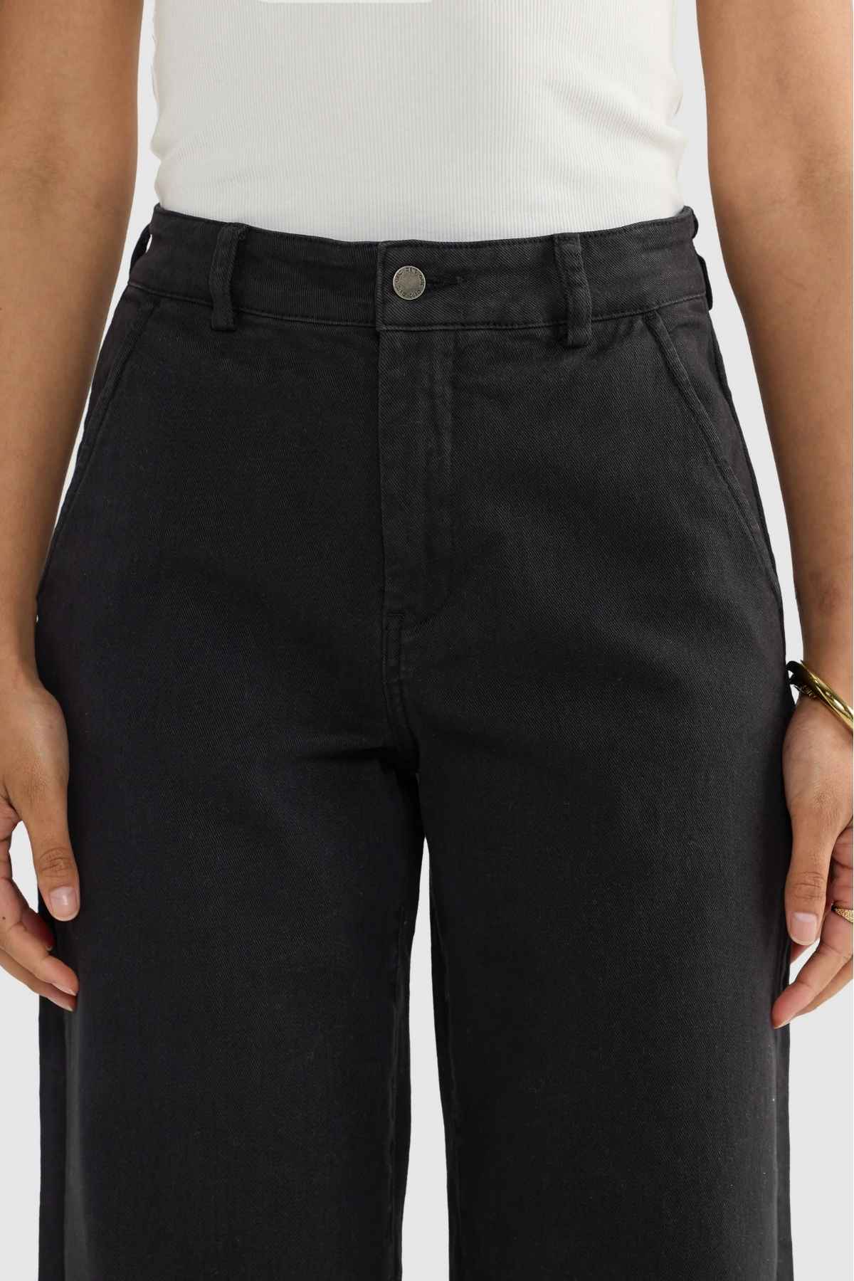 A person stands confidently, their arms relaxed at their sides, dressed in high-waisted ORTC Lottie Relaxed Twill Pants in black paired with a crisp white sleeveless top. The spotlight is on the relaxed fit and stretch cotton twill fabric, showcasing the meticulous detailing around the waist and pockets.