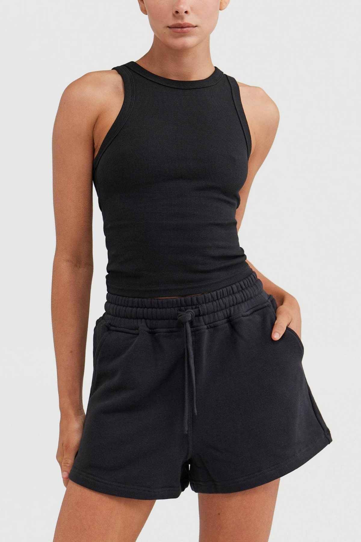 A person dressed in the ORTC Mimi Rib Tank, a black sleeveless top made from ribbed knit fabric, paired with black drawstring shorts stands against a plain background. This versatile piece is perfect for layering, with one hand casually resting in their pocket.