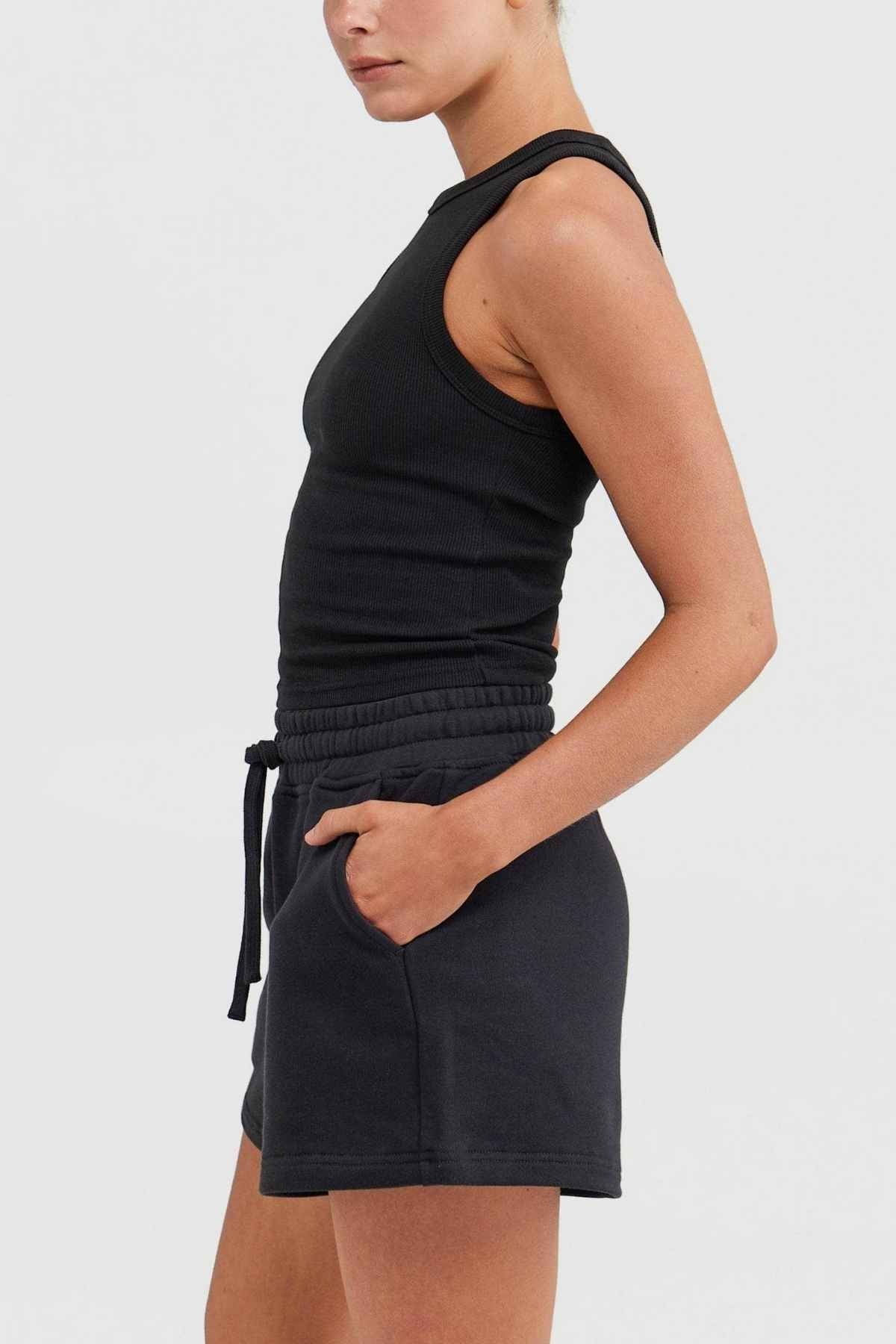 Dressed in a black ORTC Mimi Rib Tank paired with black shorts, the individual stands with hands in pockets. This tank, made from ribbed knit fabric, serves as an ideal layering piece against the simple light gray backdrop.