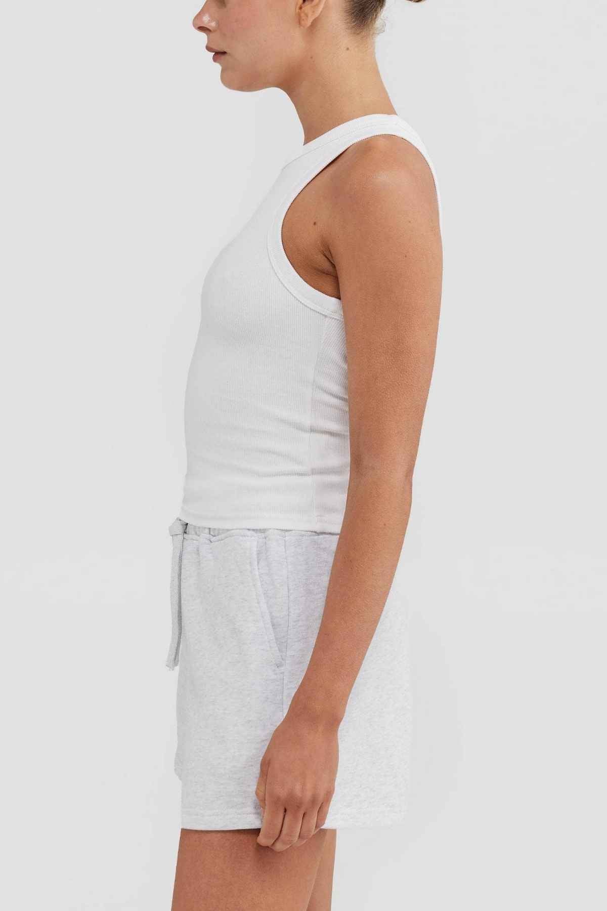 A side view reveals a person donning the ORTC Mimi Rib Tank in white, crafted from sleeveless ribbed knit fabric, paired with light gray shorts against a simple backdrop. Their hair is neatly pulled back, highlighting its versatility as an excellent layering piece.