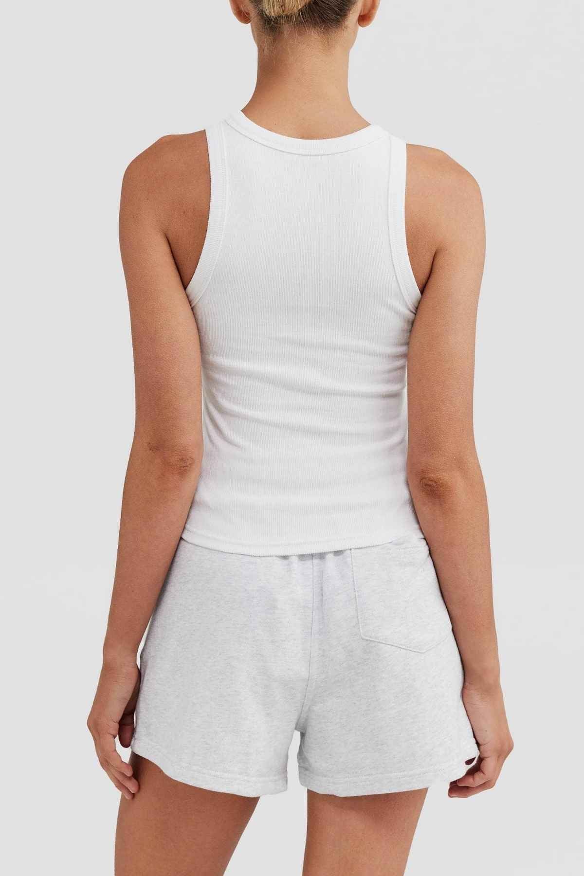A person is seen from behind wearing the ORTC Mimi Rib Tank, which features a sleeveless design in white, paired with light gray shorts against a simple light gray backdrop. The textured ribbed knit fabric makes it perfect for layering with various outfits.