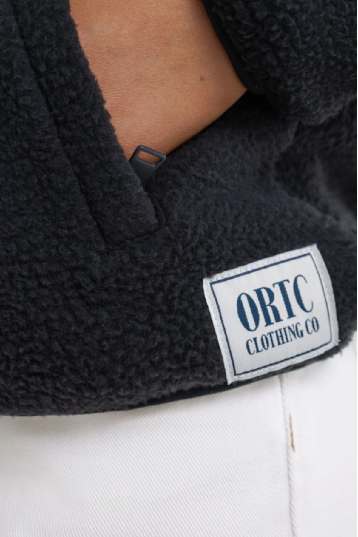 Close-up of a person wearing the ORTC Sherpa Fleece Quarter Zip in black, made from recycled polyester and featuring an "ORTC CLOTHING CO" label, styled with white pants.