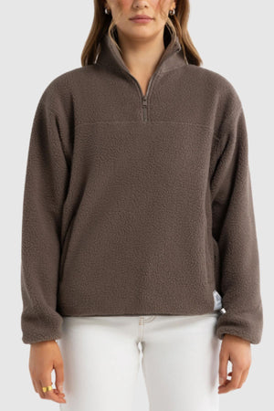 A person wearing the ORTC Sherpa Fleece Quarter Zip in brown with long sleeves stands against a plain light background, paired with white pants and gold rings.
