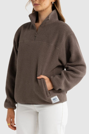 A person wears the ORTC Sherpa Fleece Quarter Zip in brown, featuring a small rectangular patch near the hem, one hand in the front pocket, paired with white pants. This cozy top is made from recycled polyester for sustainability.