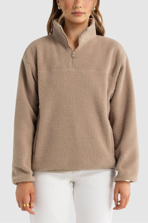 A woman models the ORTC Sherpa Fleece Quarter Zip, a beige pullover made from recycled polyester, paired with white pants against a plain light gray background. Her face is partially visible and her hands are relaxed at her sides.