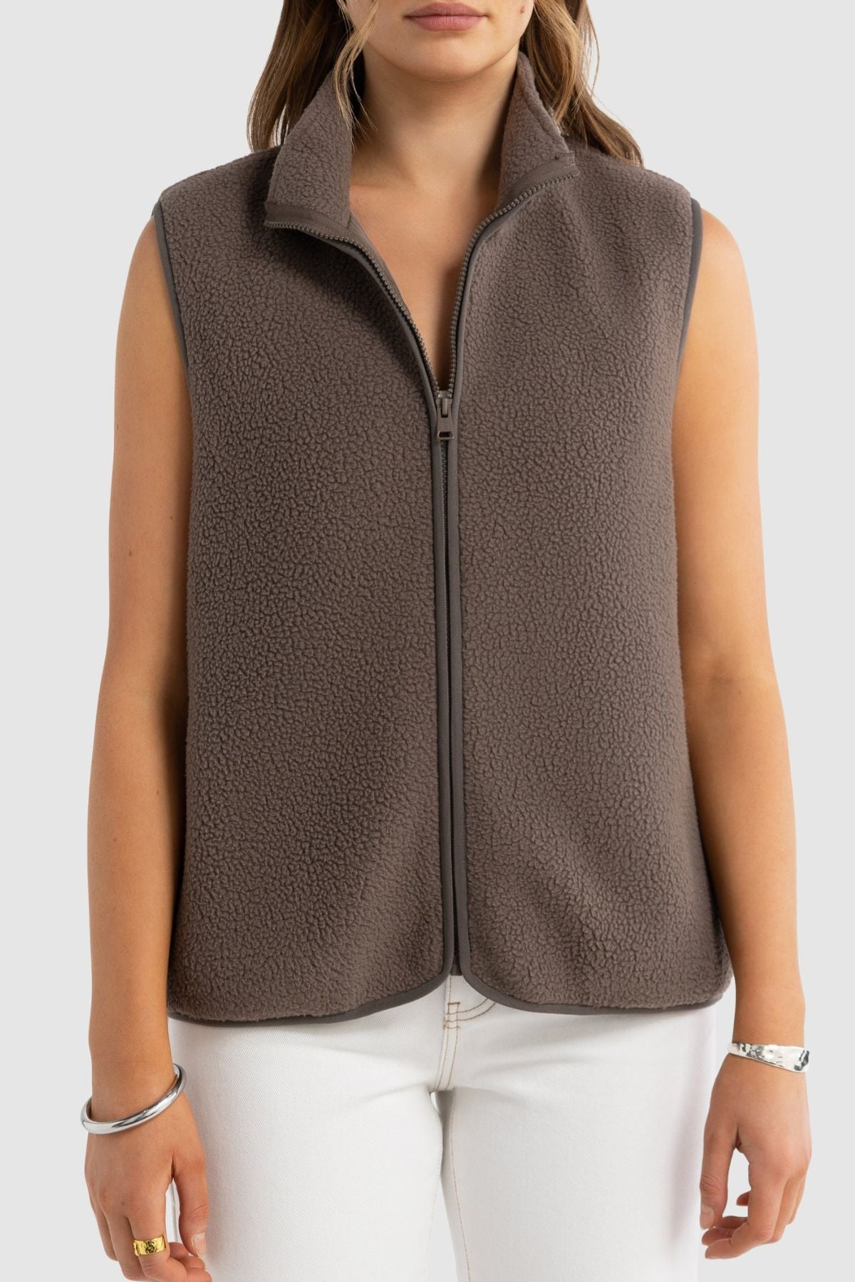 Wearing the ORTC Sully Sherpa Fleece Vest—a sleeveless, zip-up high-collar vest—paired with white pants and minimal jewelry against a plain background makes for stylish, versatile layering.