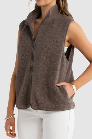 A woman models the ORTC Sully Sherpa Fleece Vest—a sleeveless, brown vest made from recycled polyester—layered over a white top and pants, with one hand in her pocket, set against a light gray background.
