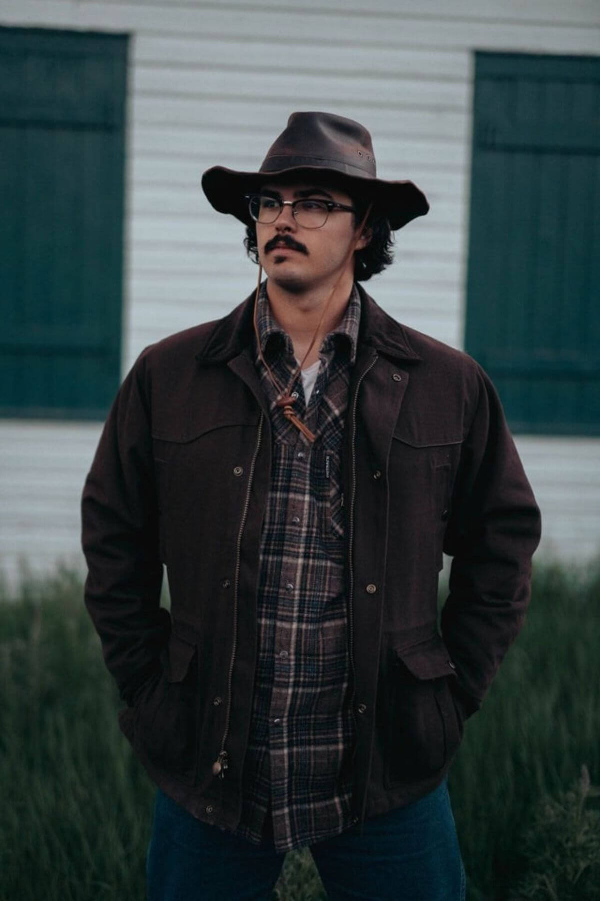 A man with a mustache, wearing glasses and a dark cowboy hat, stands in front of a white building with green shutters. He is dressed in an Outback Cattleman Jacket over a plaid shirt, his hands nestled in the jacket's multiple pockets, looking off into the distance like an outdoor companion ready for adventure.