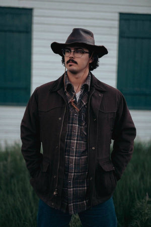 A man with a mustache, wearing glasses and a dark cowboy hat, stands in front of a white building with green shutters. He is dressed in an Outback Cattleman Jacket over a plaid shirt, his hands nestled in the jacket's multiple pockets, looking off into the distance like an outdoor companion ready for adventure.