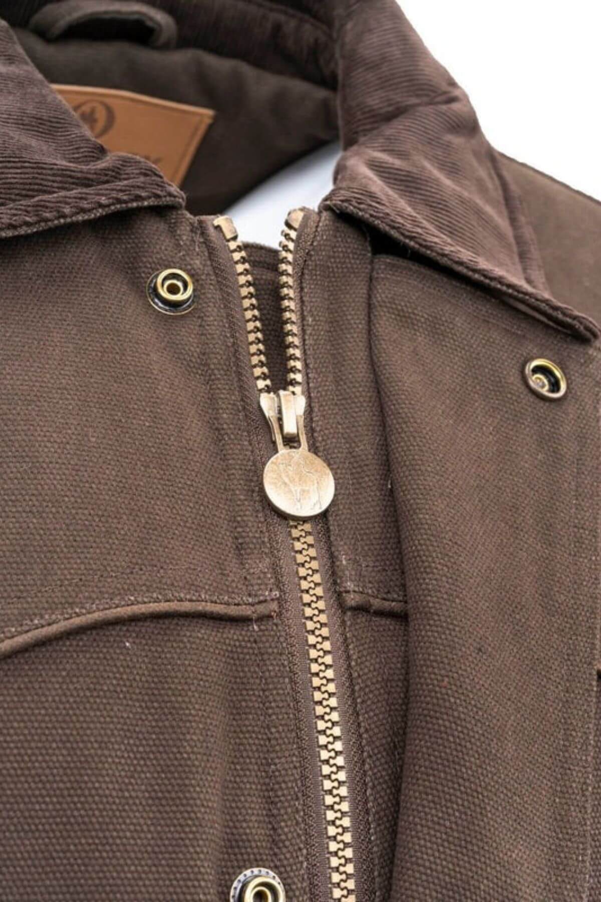 Close-up of the Outback Cattleman Jacket showcases a metal zipper partially zipped, featuring a circular design on the zipper pull. The jacket boasts multiple pockets with flap closures and brass snap buttons. The inner collar is lined with corduroy material, making it an ideal companion for outdoor adventures.