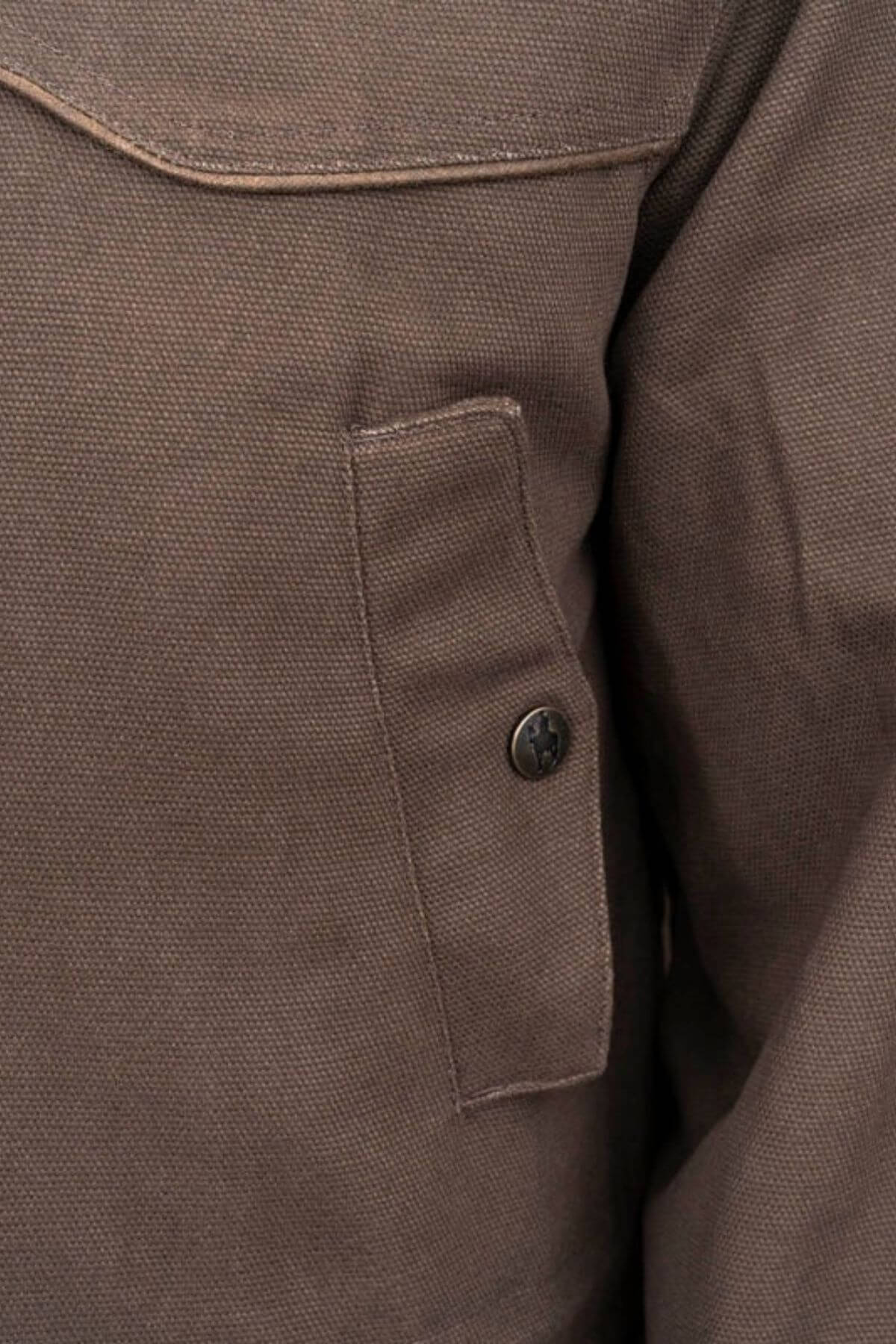 Close-up of the textured brown fabric of an Outback Cattleman Jacket, highlighting a buttoned pocket with visible stitching. The material looks thick and durable. Featuring a brass 2-way zipper, this jacket offers both style and functionality for outdoor use.