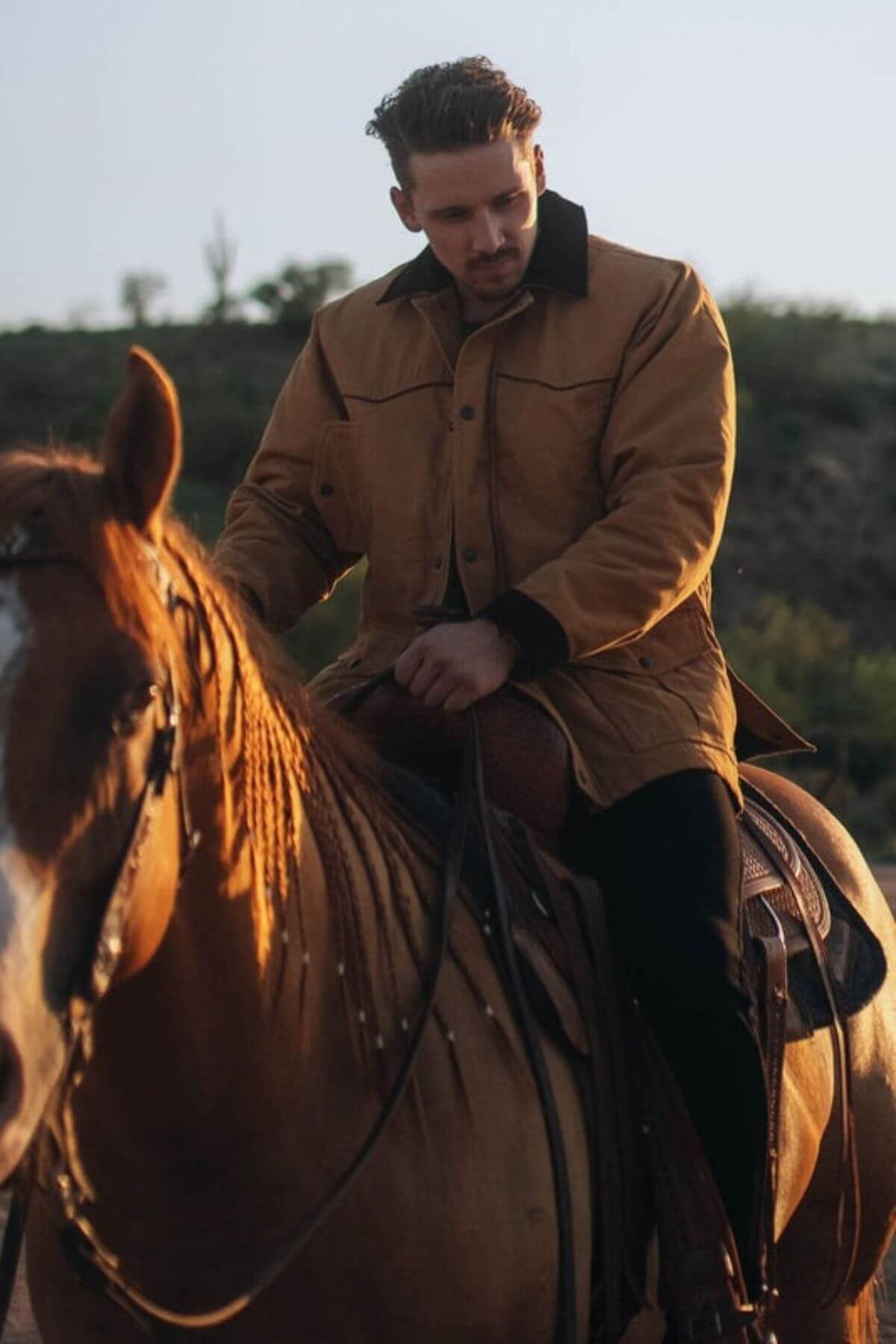 A man in an Outback Cattleman Jacket rides a brown horse outdoors, sunlight highlighting them with shrubs and a hill in the background.
