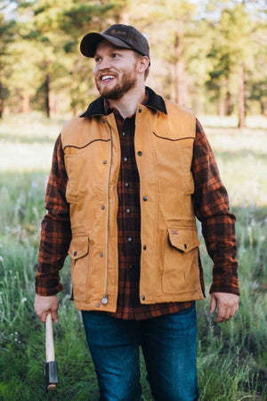 A man with a beard, wearing a brown cap, brown checkered shirt, and an Outback Cattleman Vest made of machine washable cotton fabric, stands outdoors in a grassy area holding an axe. He is smiling and looking to his left while tall trees can be seen in the background.