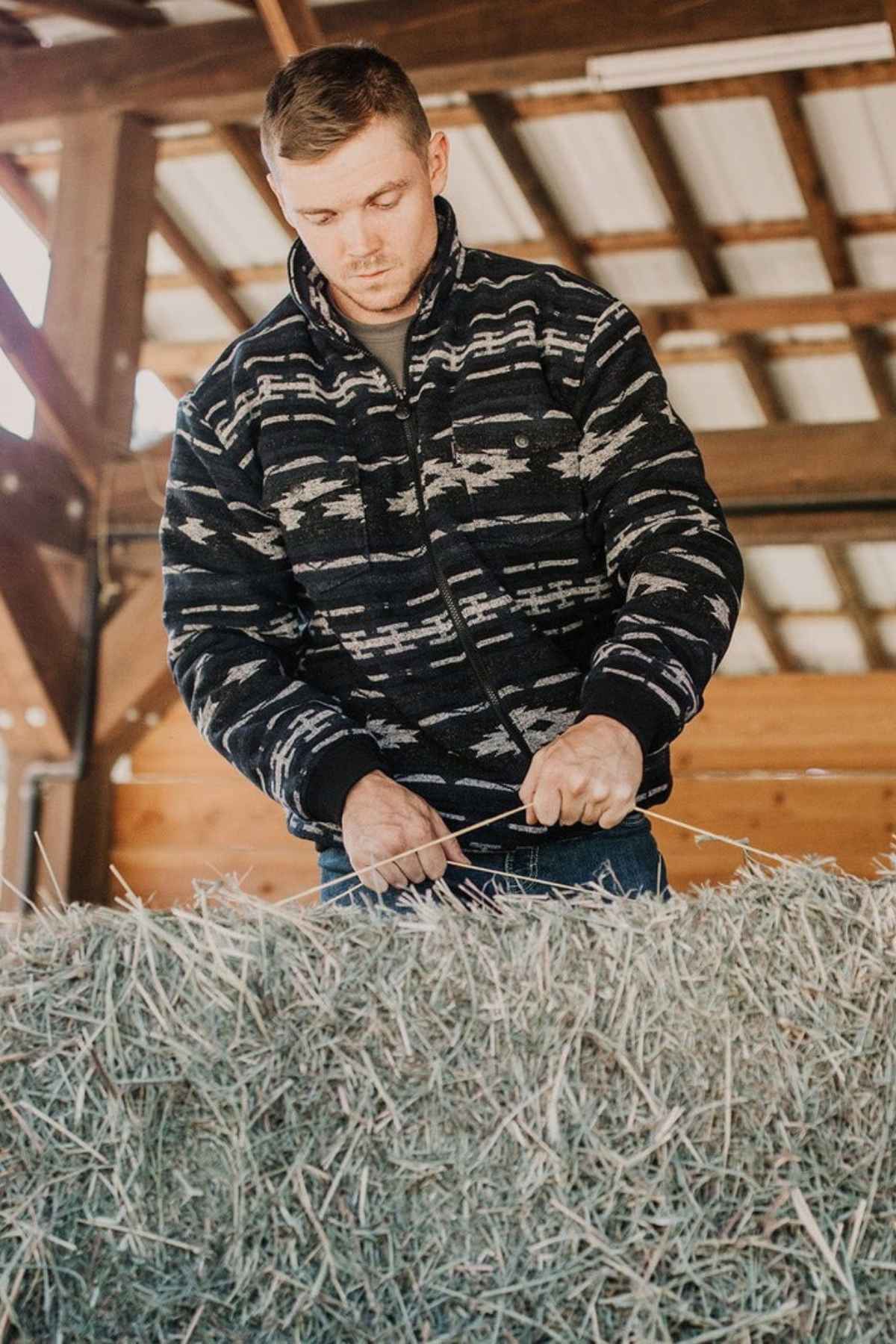 A man wearing an Outback Marc Jacket with a 2-way zipper ties twine around a hay bale inside a wood structure with a metal roof, focused on his task while hay is visible in the foreground.