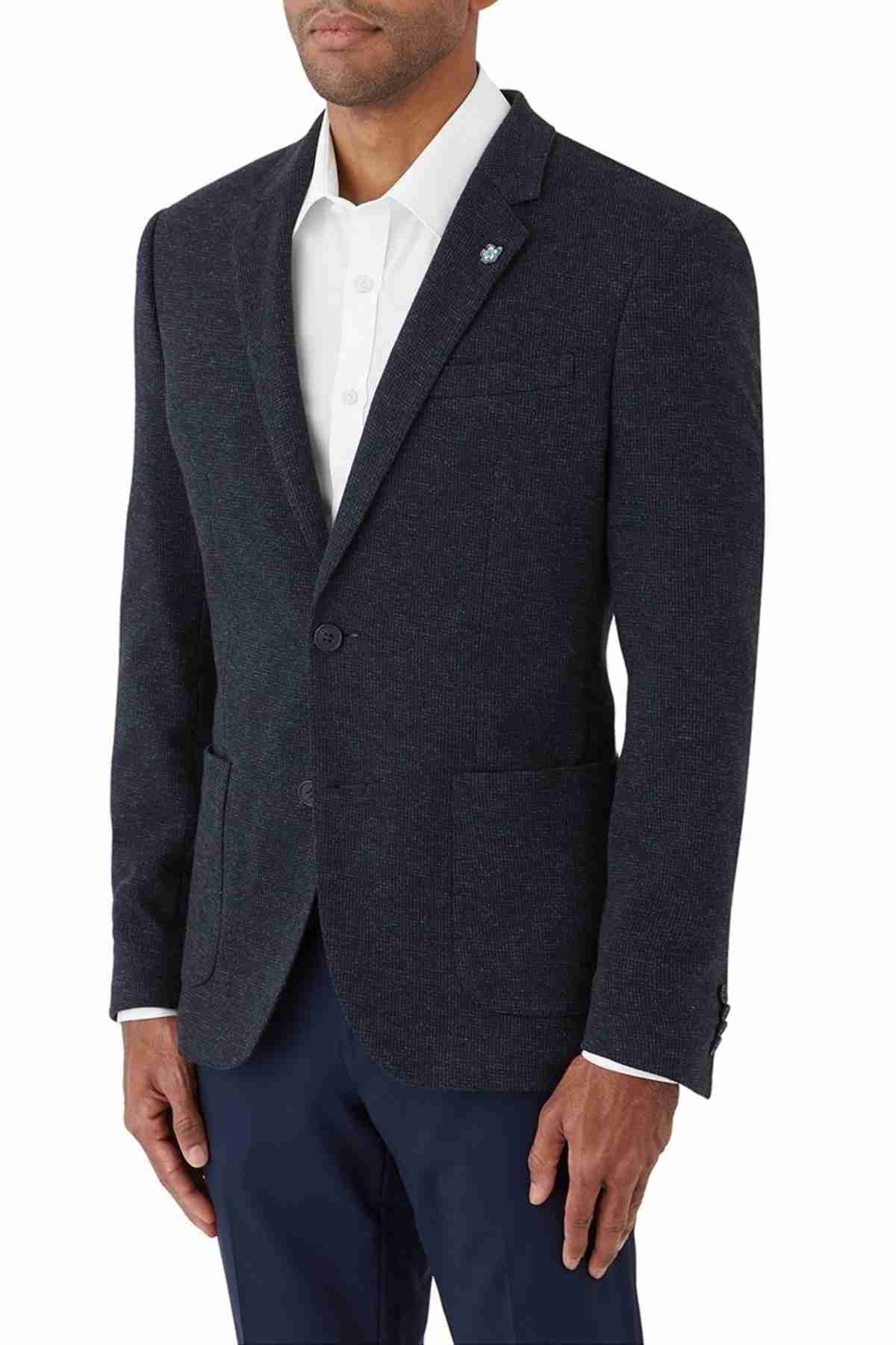 A man wearing the Uberstone Zeller Jacket, a dark blue wool-blend blazer with a subtle pattern. It's buttoned in the front and has a small pin on the lapel, paired with a white dress shirt and skinny fit dark pants.