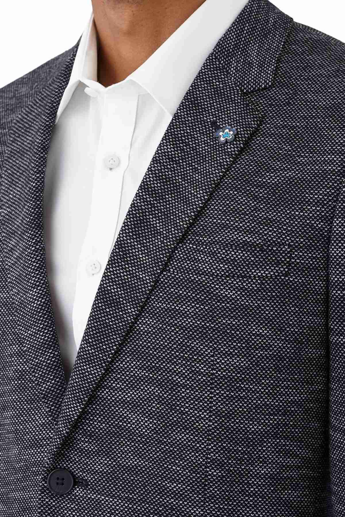 A person wears the Uberstone Zeller Jacket, a textured navy wool blend blazer with a notched lapel and small blue flower pin, layered over a white dress shirt. The image focuses on the upper torso, without showing the face.