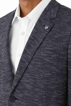 A person wears the Uberstone Zeller Jacket, a textured navy wool blend blazer with a notched lapel and small blue flower pin, layered over a white dress shirt. The image focuses on the upper torso, without showing the face.