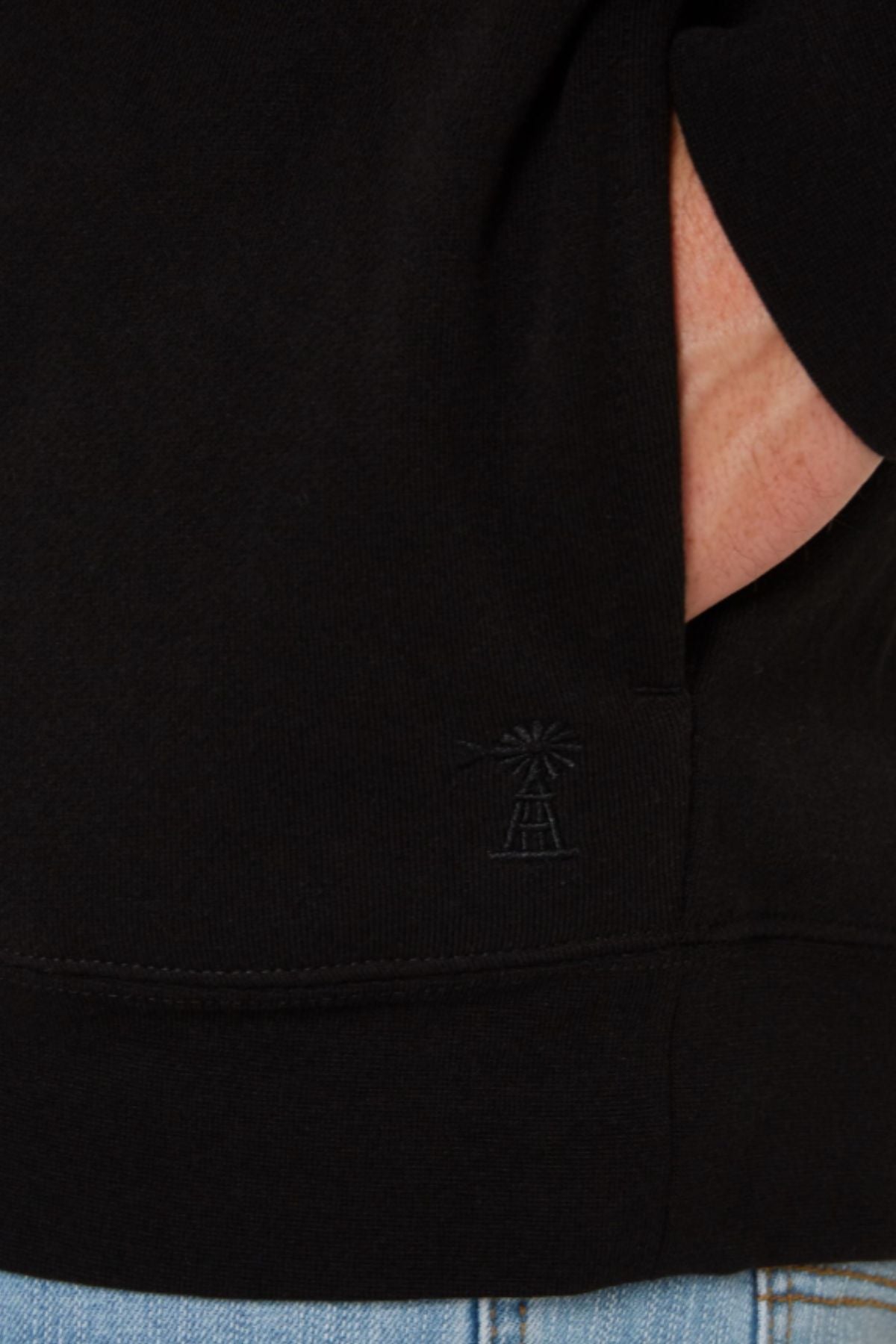 Close-up of a person wearing a black Pilbara Classic Fleece Pullover, featuring a small embossed windmill design near the hem. The hand rests partly in a pocket, paired with light blue denim jeans.