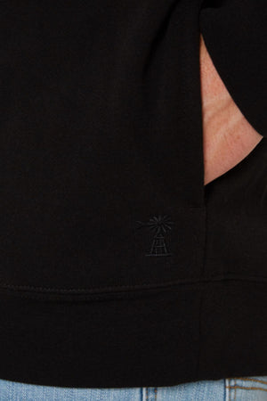 Close-up of a person wearing a black Pilbara Classic Fleece Pullover, featuring a small embossed windmill design near the hem. The hand rests partly in a pocket, paired with light blue denim jeans.